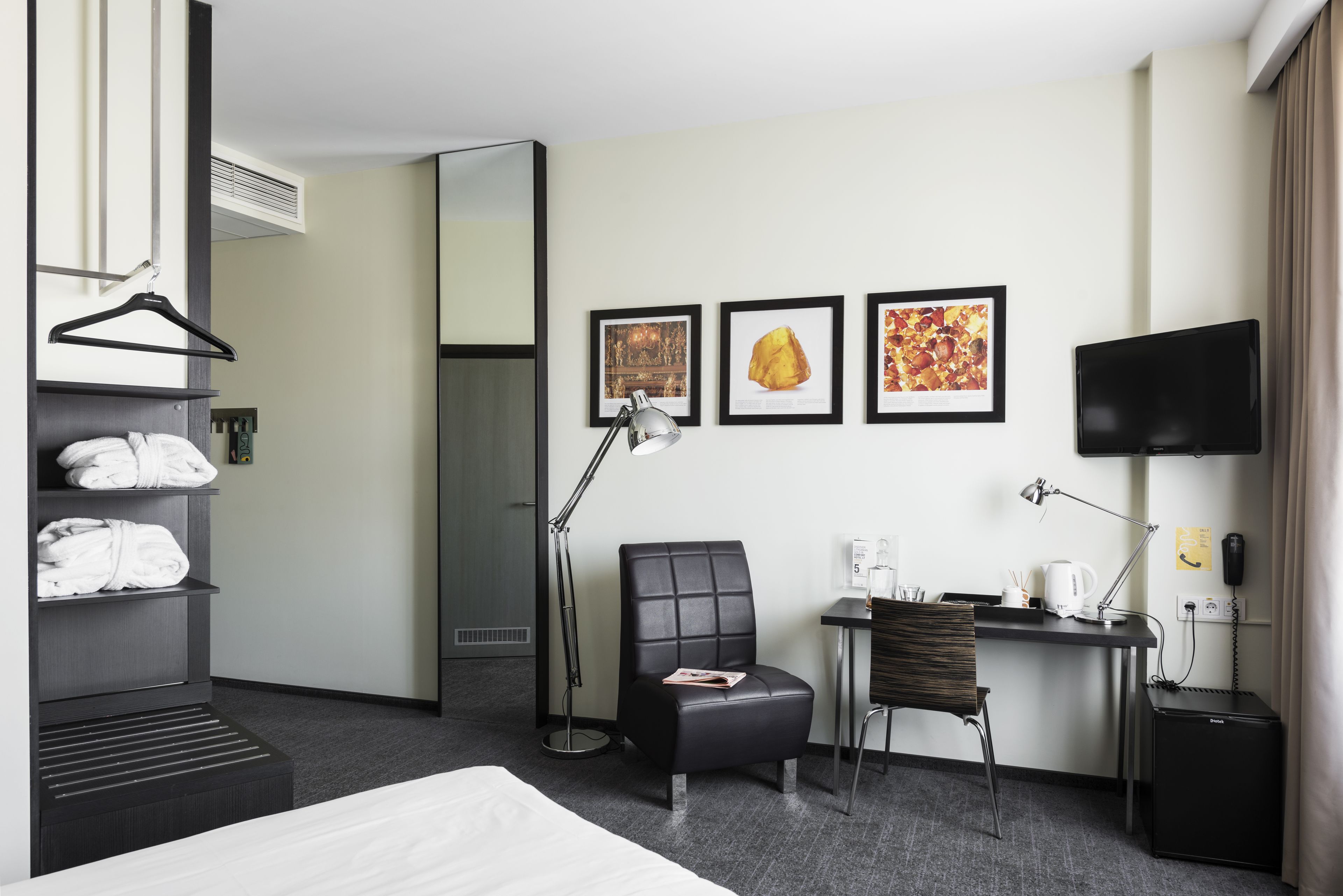 A hotel room with a bed, a desk with a chair, a black leather chair, a black TV, a desk lamp, a mirror, and a wall-mounted shelf with towels.