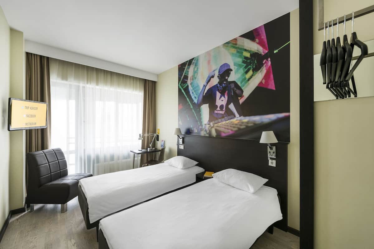 A hotel room with two beds, a desk, a chair, a TV, and a large abstract painting on the wall.