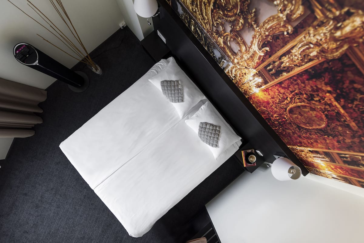 A neatly made bed with white linens and a gray pillow, a wall-mounted lamp, and a decorative wall with a golden design.
