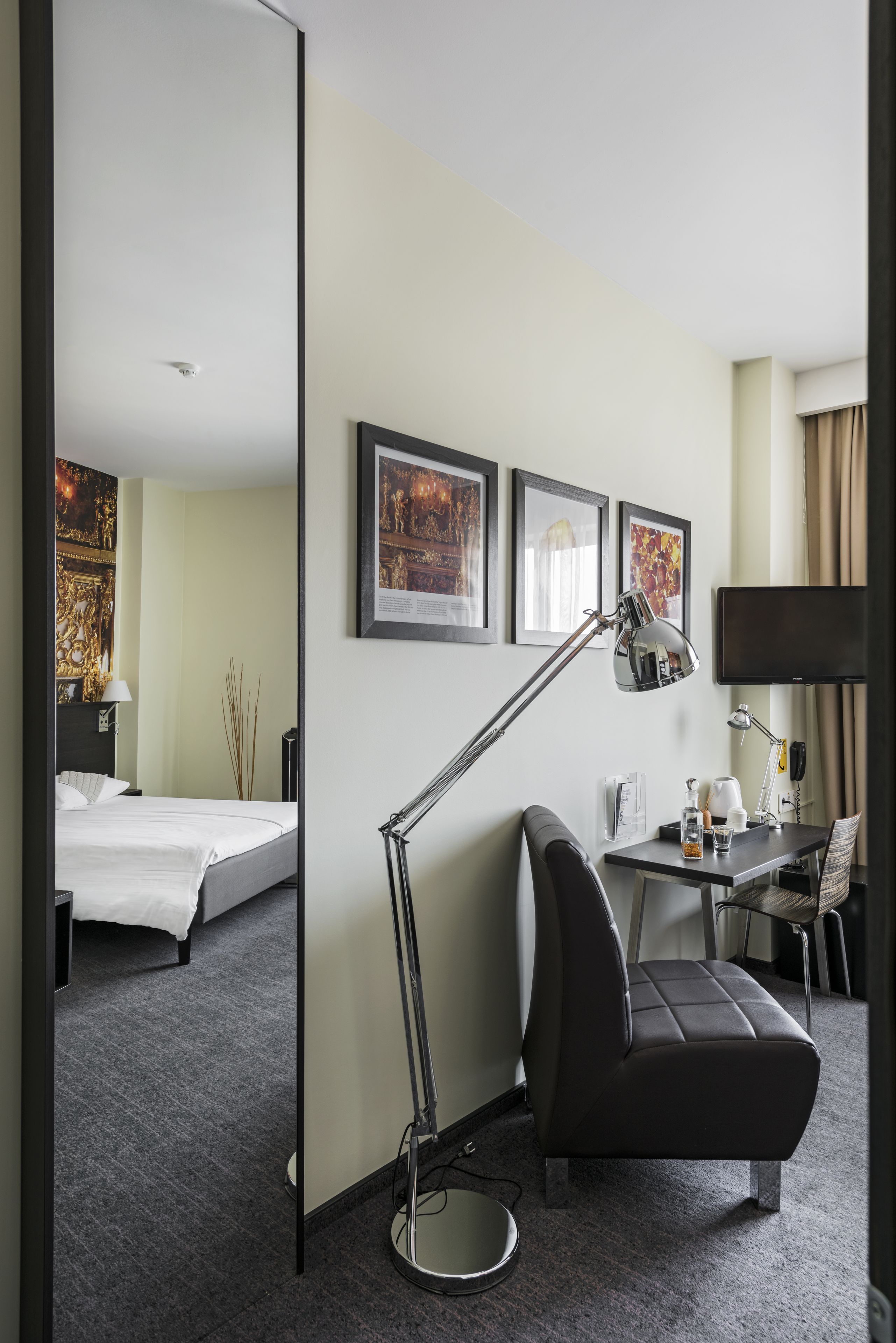 A hotel room with a bed, a desk, a chair, a standing lamp, and framed artwork on the wall.