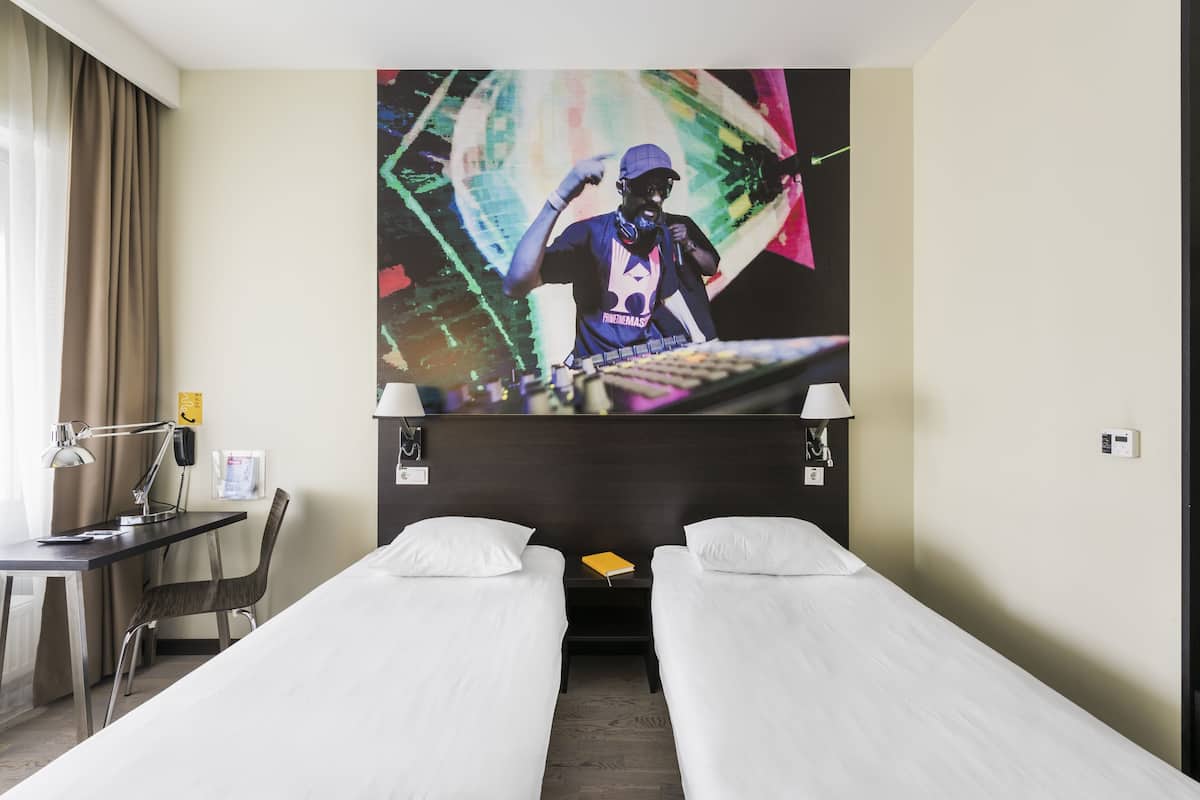 A hotel room with two single beds, a desk with a chair, a lamp, a phone, and a wall-mounted TV displaying a DJ.