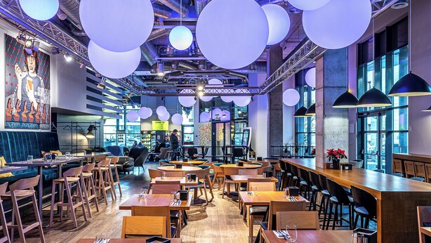 Un restaurante espacioso con techos altos, conductos expuestos y lámparas esféricas colgantes. Las mesas están dispuestas en línea con sillas de madera.