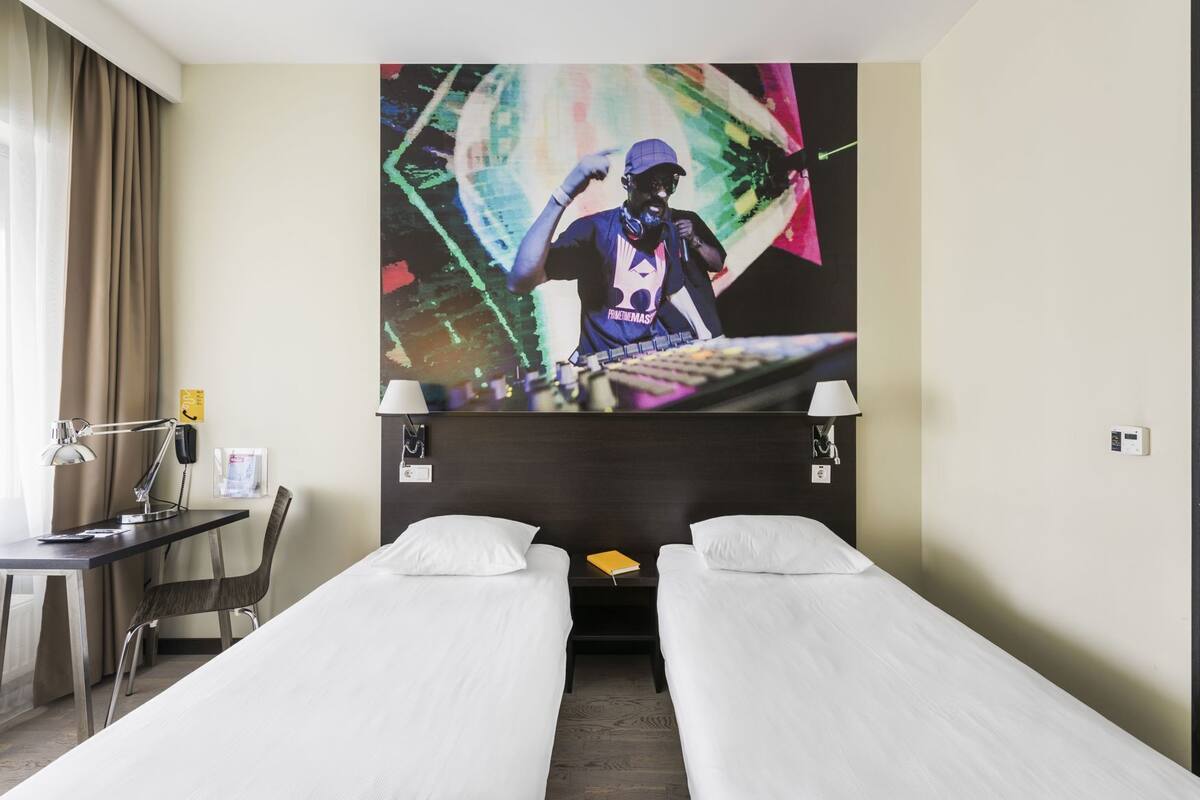 A hotel room with two single beds, a desk with a chair, a lamp, a phone, and a large abstract painting on the wall.