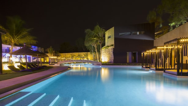 A swimming pool with a stone bridge, palm trees, and modern buildings at night. - The Rhino Resort Hotel & Spa (Mbour)