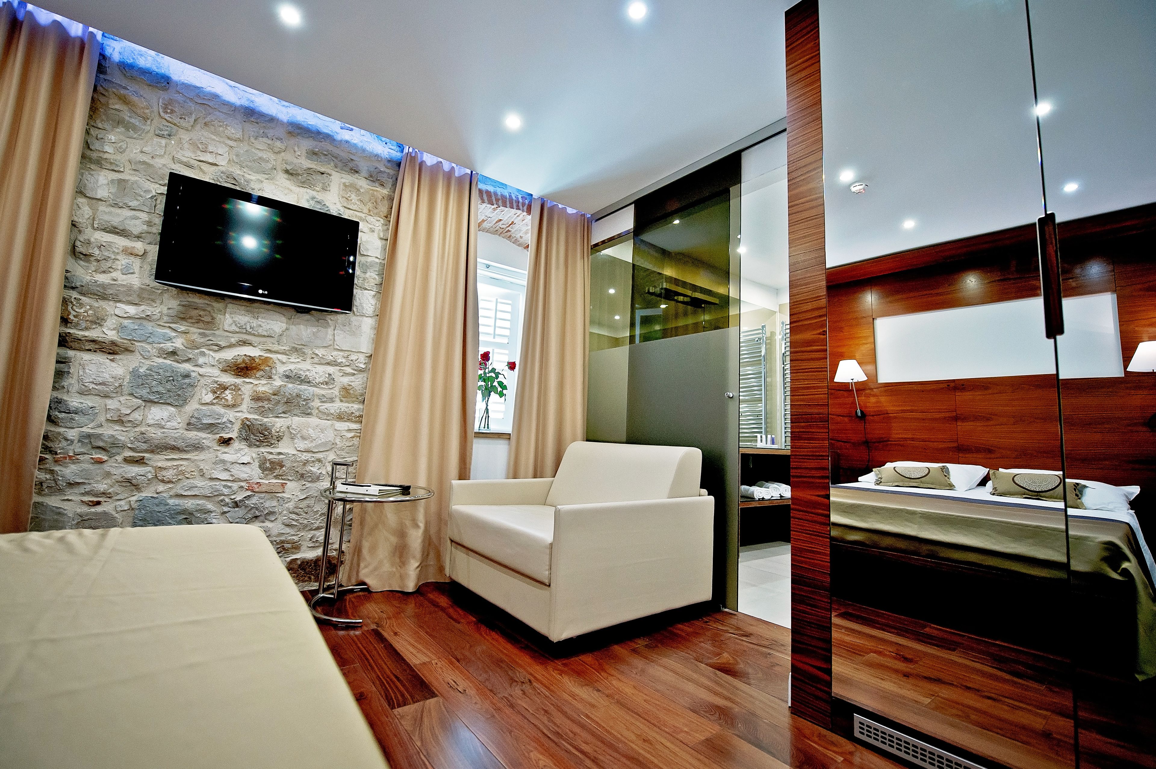 Habitación de hotel moderna con pared de piedra, televisor de pantalla plana, sillón blanco, cama con cabecero de madera y una mesita de noche.