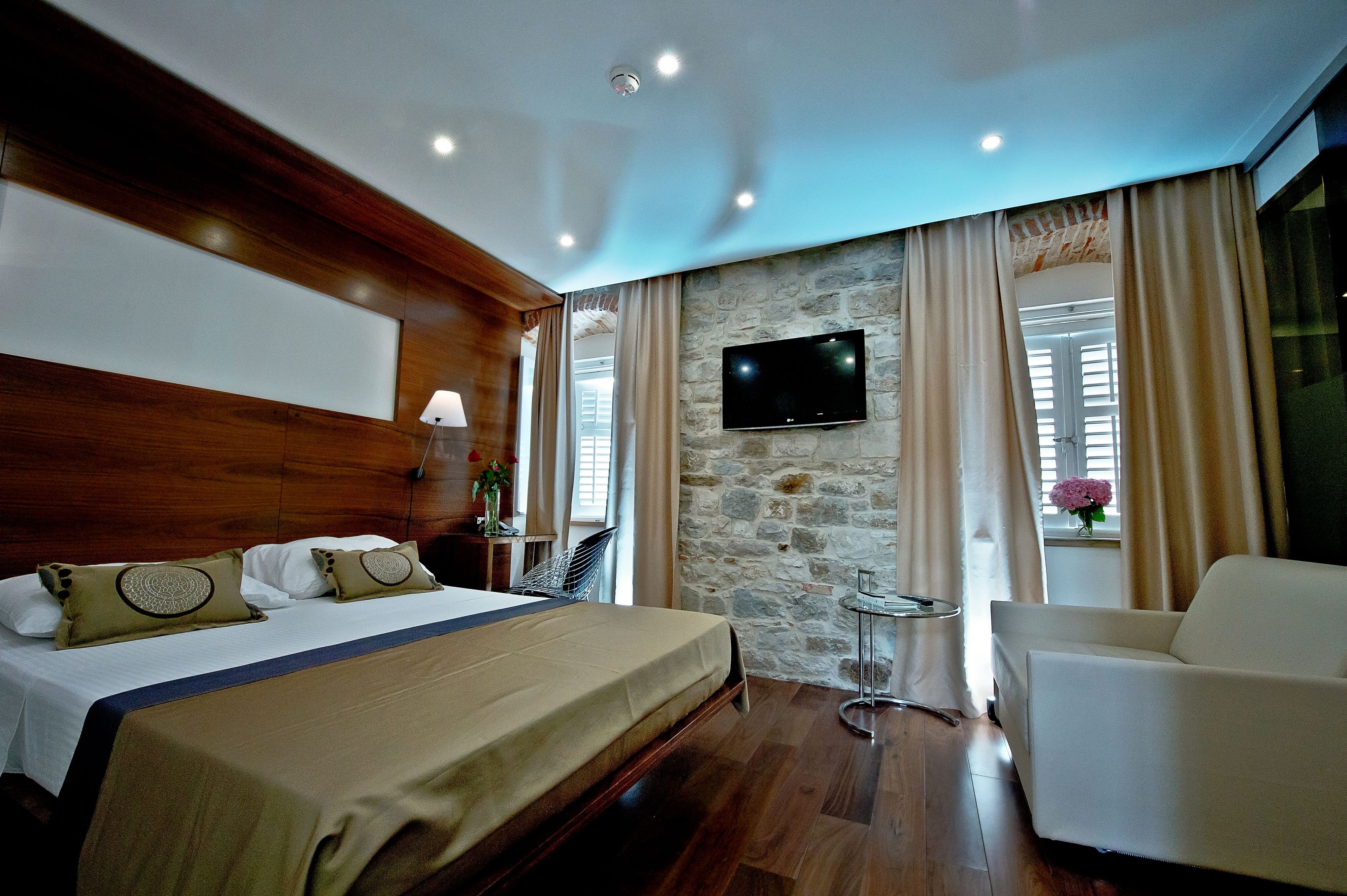 A hotel room with a bed, a TV mounted on a stone wall, a chair, and a window with curtains.