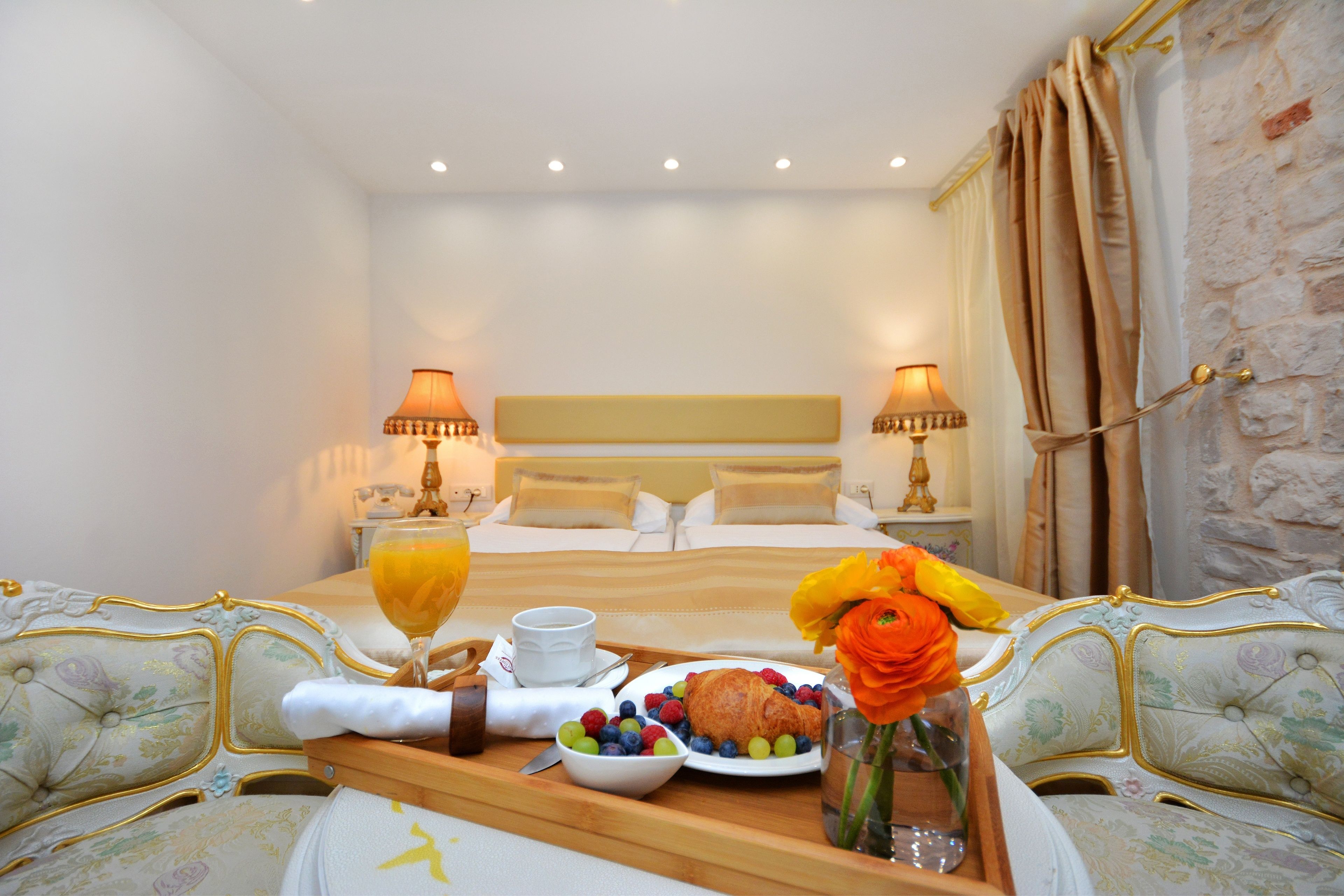 A hotel room with a bed, bedside lamps, a breakfast tray with fruit, a vase with flowers, and a stone wall.