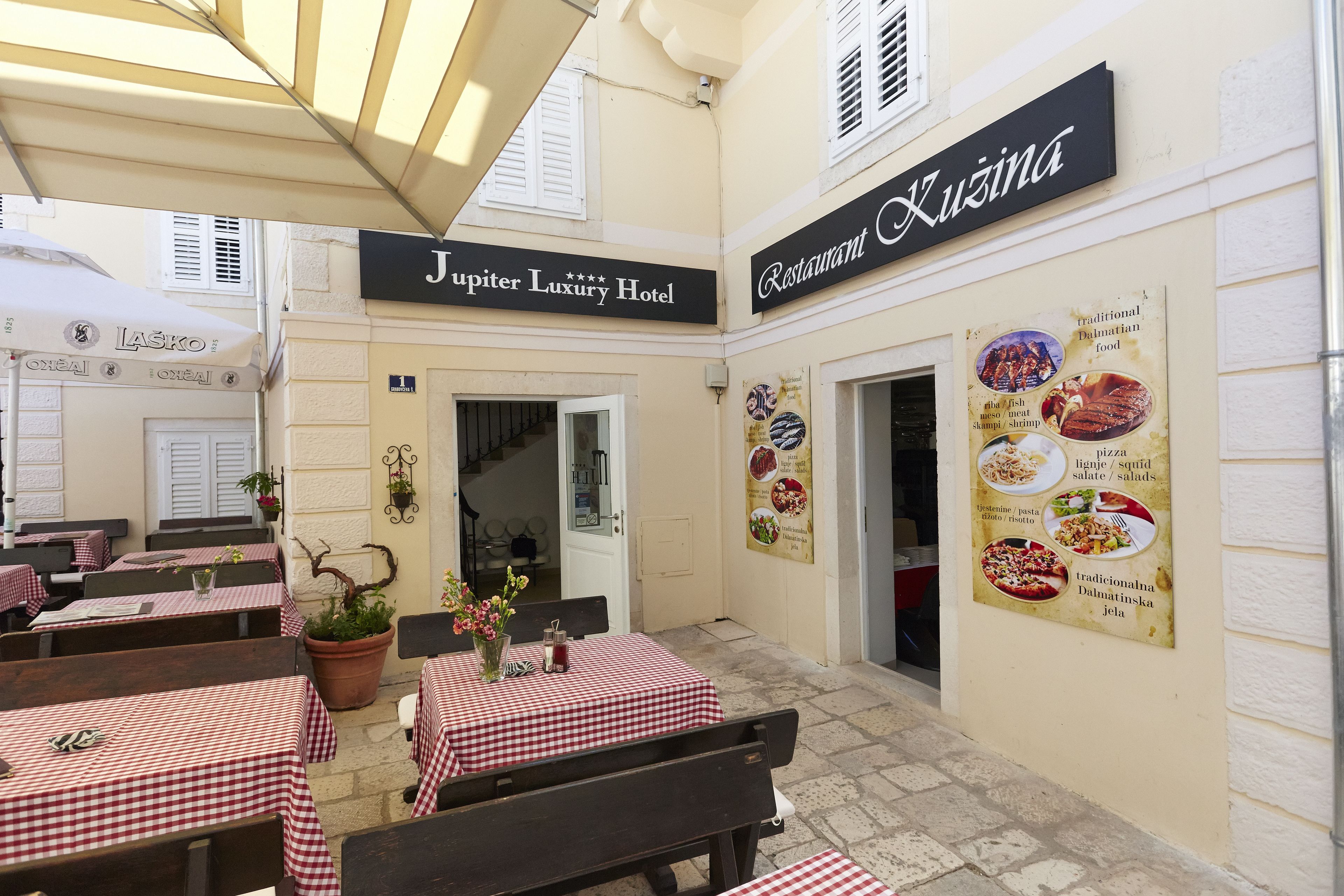 An outdoor dining area with checkered tables and benches, a sign for 'Jupiter Luxury Hotel', and a menu board for 'Kuzina' restaurant.