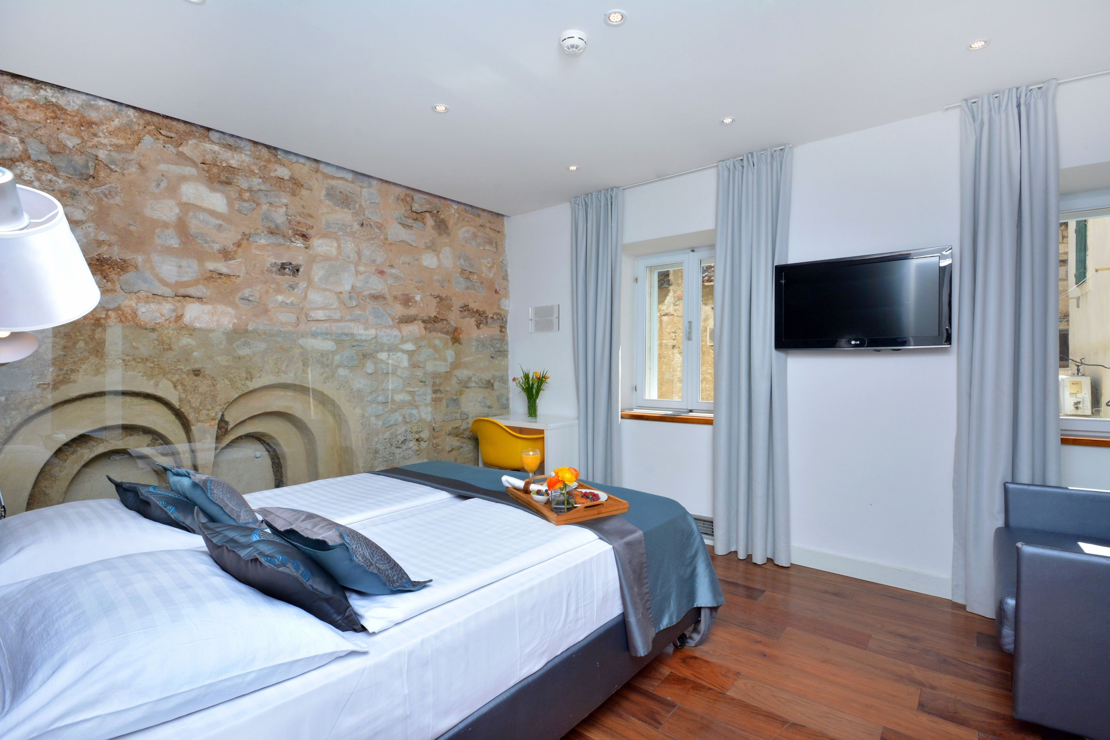 Ein modernes Hotelzimmer mit einer Wand aus Stein, einem Bett mit einem Tablett mit Frühstück, einem Flachbildfernseher und einem Fenster mit Vorhängen.
