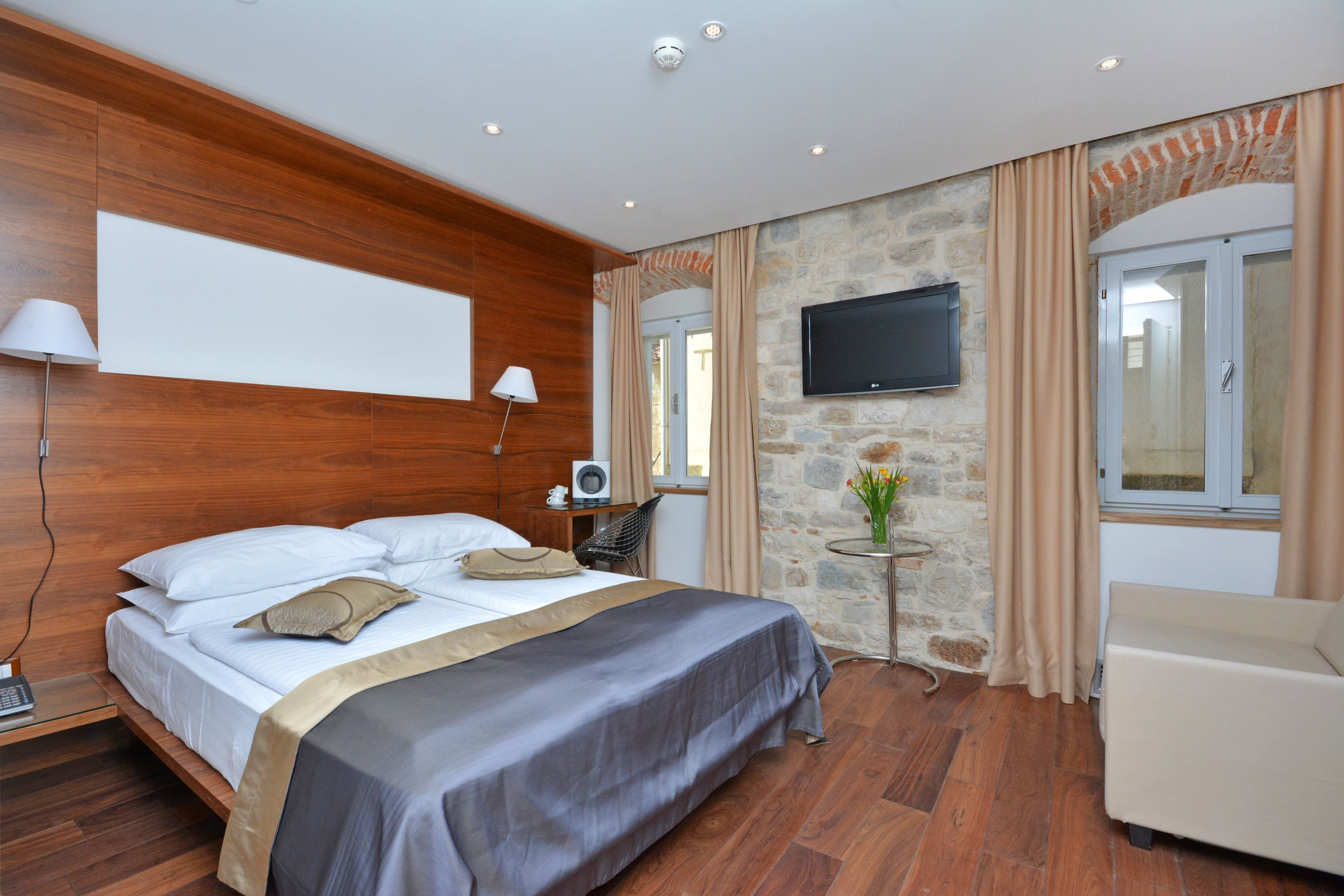 A hotel room with a wooden headboard, a bed with white linens, a flat-screen TV mounted on the wall, a small table with a vase, and a window with curtains.