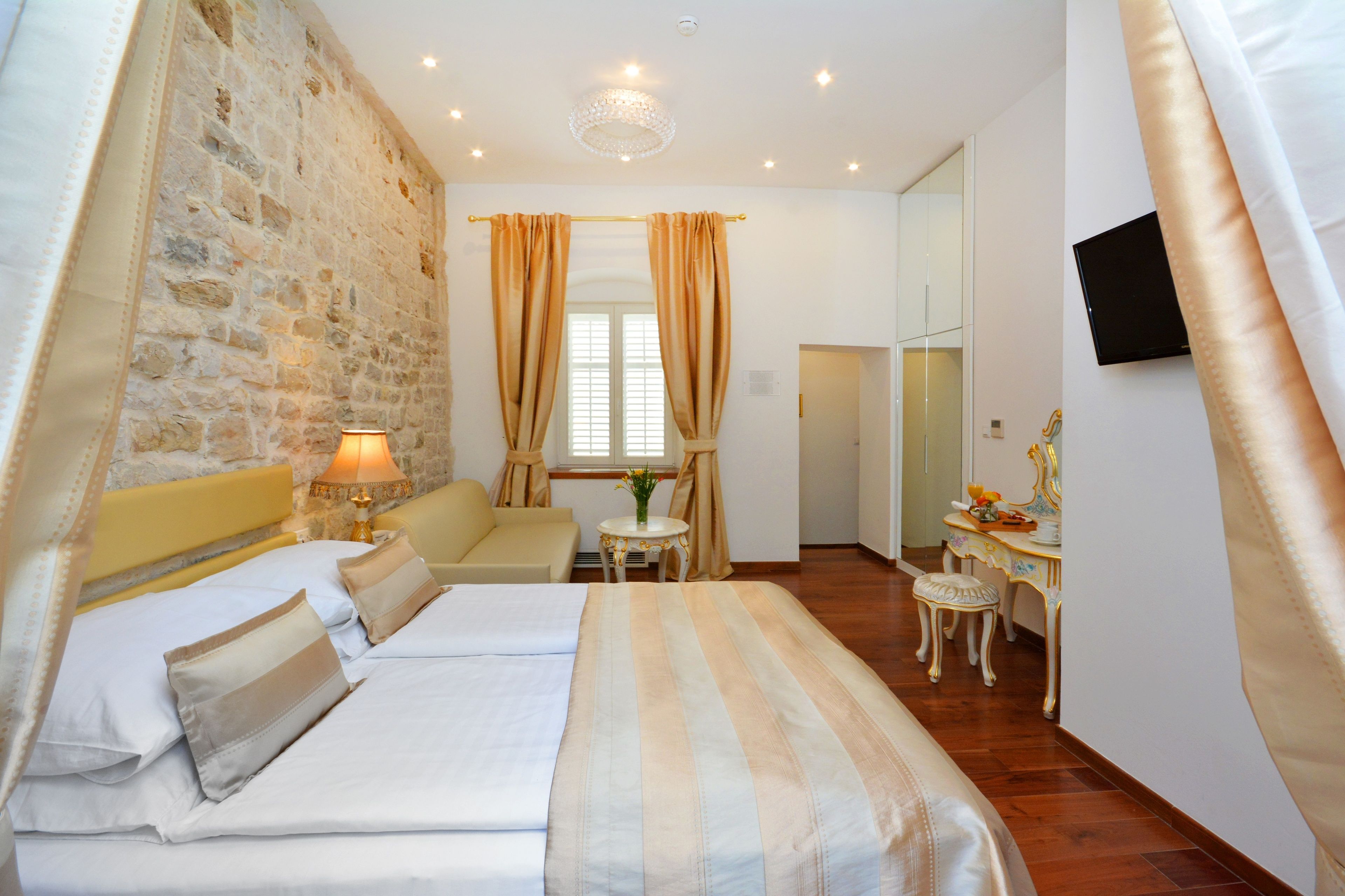 Una habitación de hotel con pared de piedra, una cama con ropa de cama a rayas, una mesita con una lámpara, una silla y un escritorio con espejo.