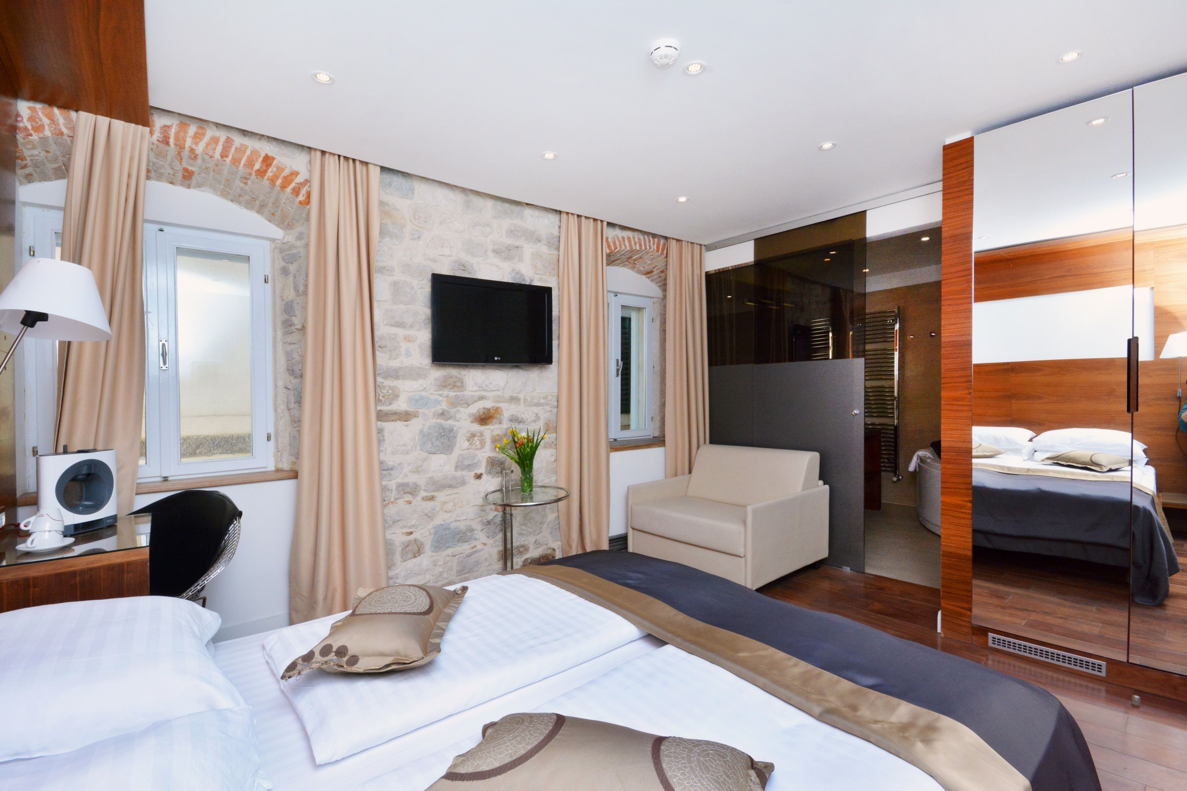 A modern hotel room with a stone accent wall, a flat-screen TV, a sofa, a bed with a wooden headboard, and a mirror cabinet.