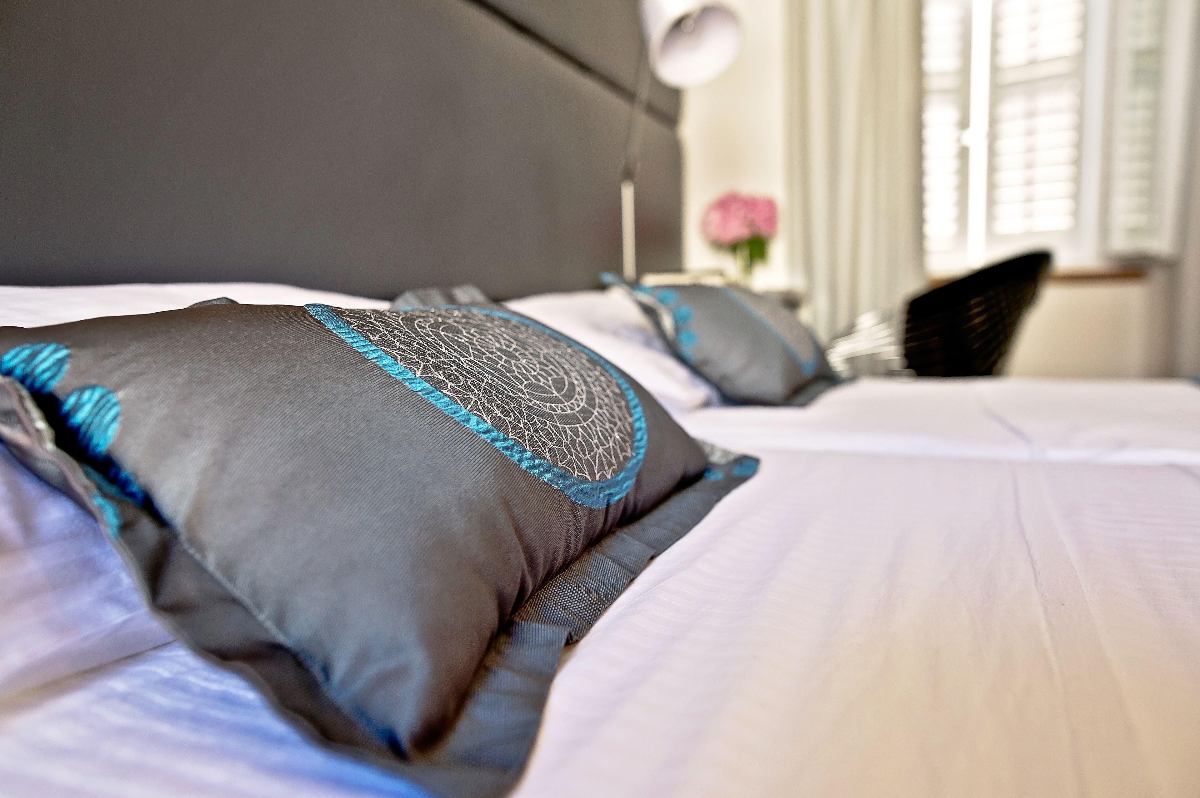 Ein ordentlich bezogenes Bett mit grauen und blauen Kissen, einem dunklen Kopfteil und einem Nachttisch mit Lampe.
