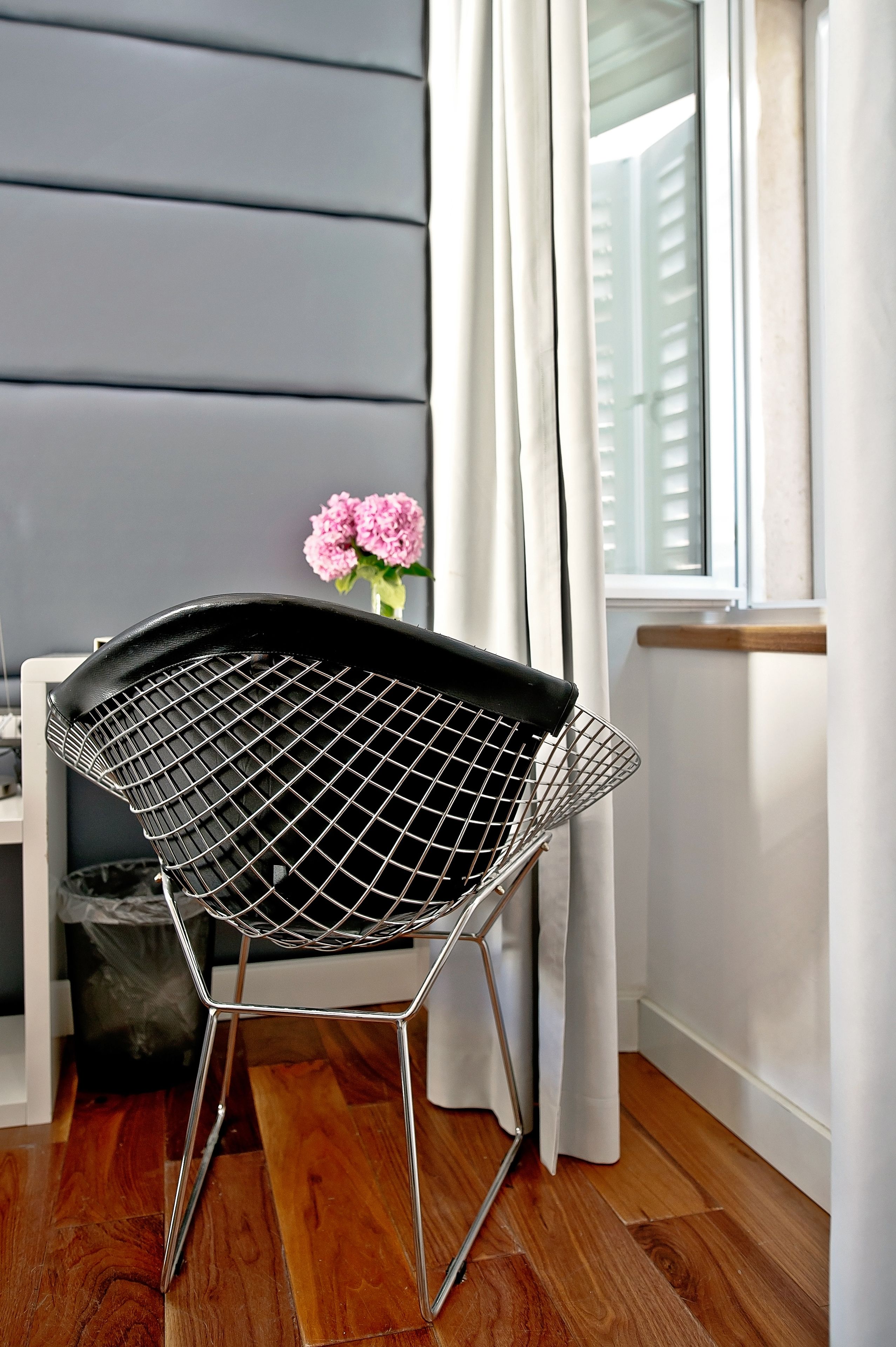 Una silla moderna con estructura metálica y asiento de malla, ubicada en una habitación con piso de madera, una ventana con cortinas blancas y un jarrón con flores rosas sobre una mesa.