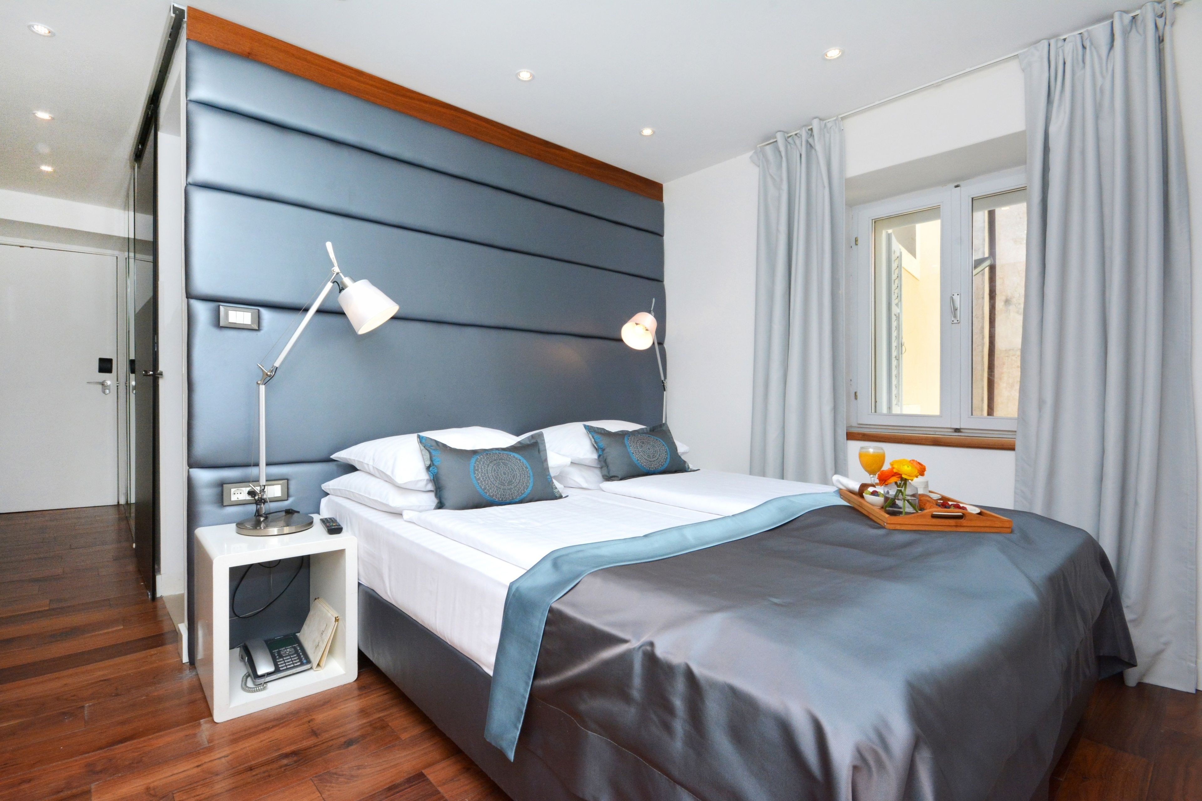 A modern hotel room with a large bed, a bedside table with a lamp, a nightstand with a phone, and a window with curtains.