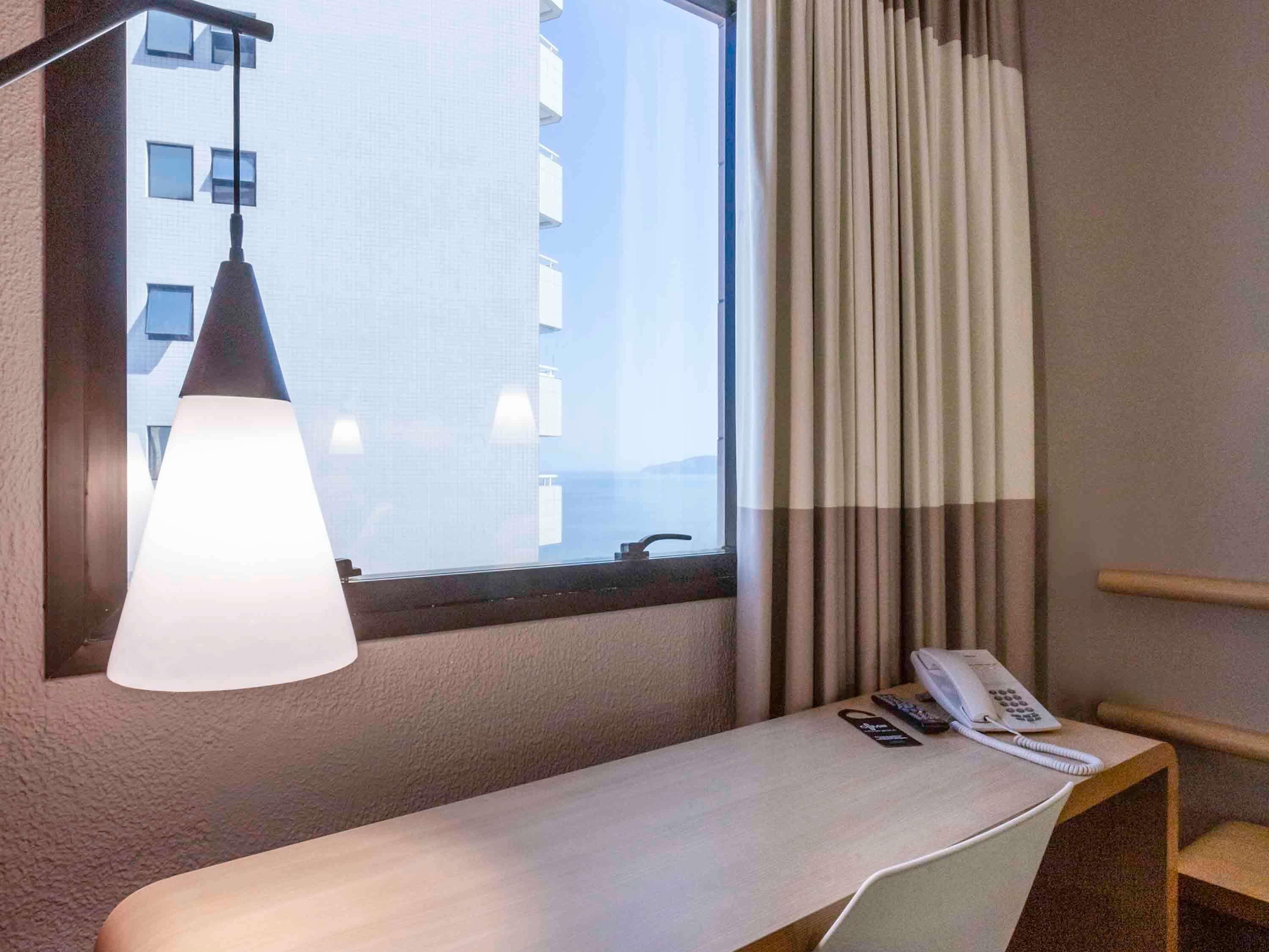 A hotel room with a bedside table, a lamp, a telephone, and a window with curtains.