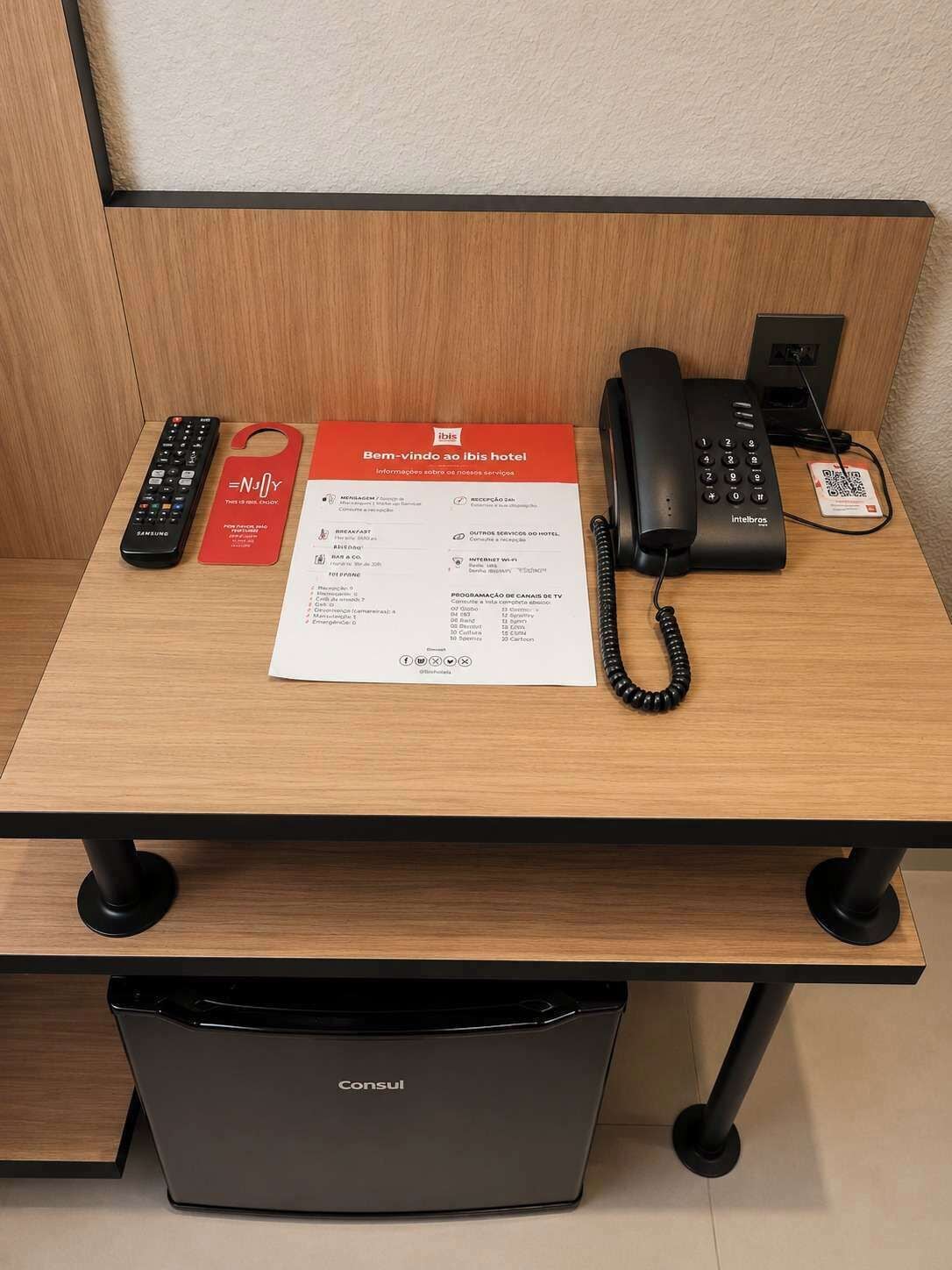 A hotel desk with a Samsung remote, a hotel brochure, a telephone, and a QR code.