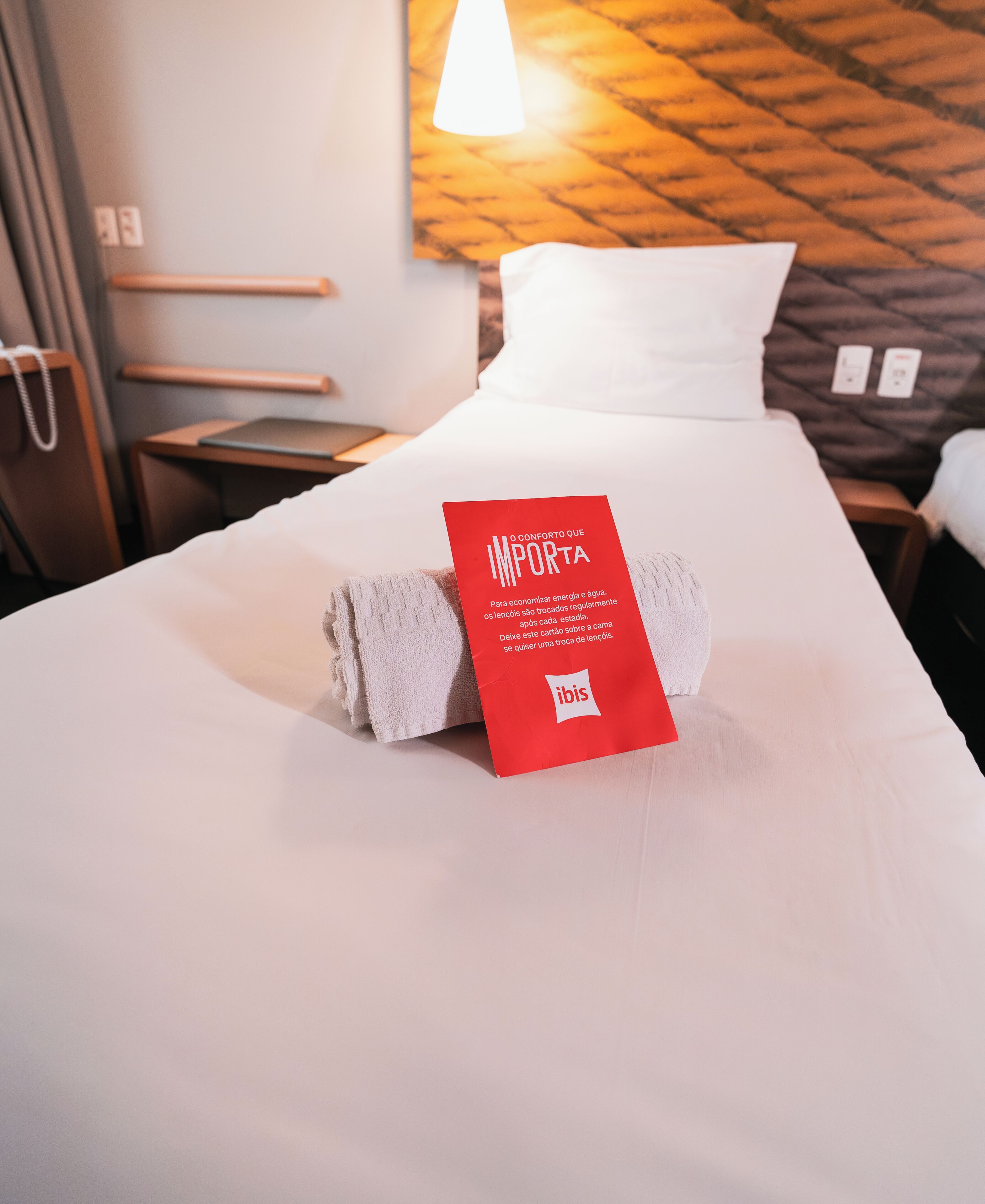 A neatly made bed with a red tag on the pillows, a wooden headboard, a bedside table, and a wall-mounted lamp.