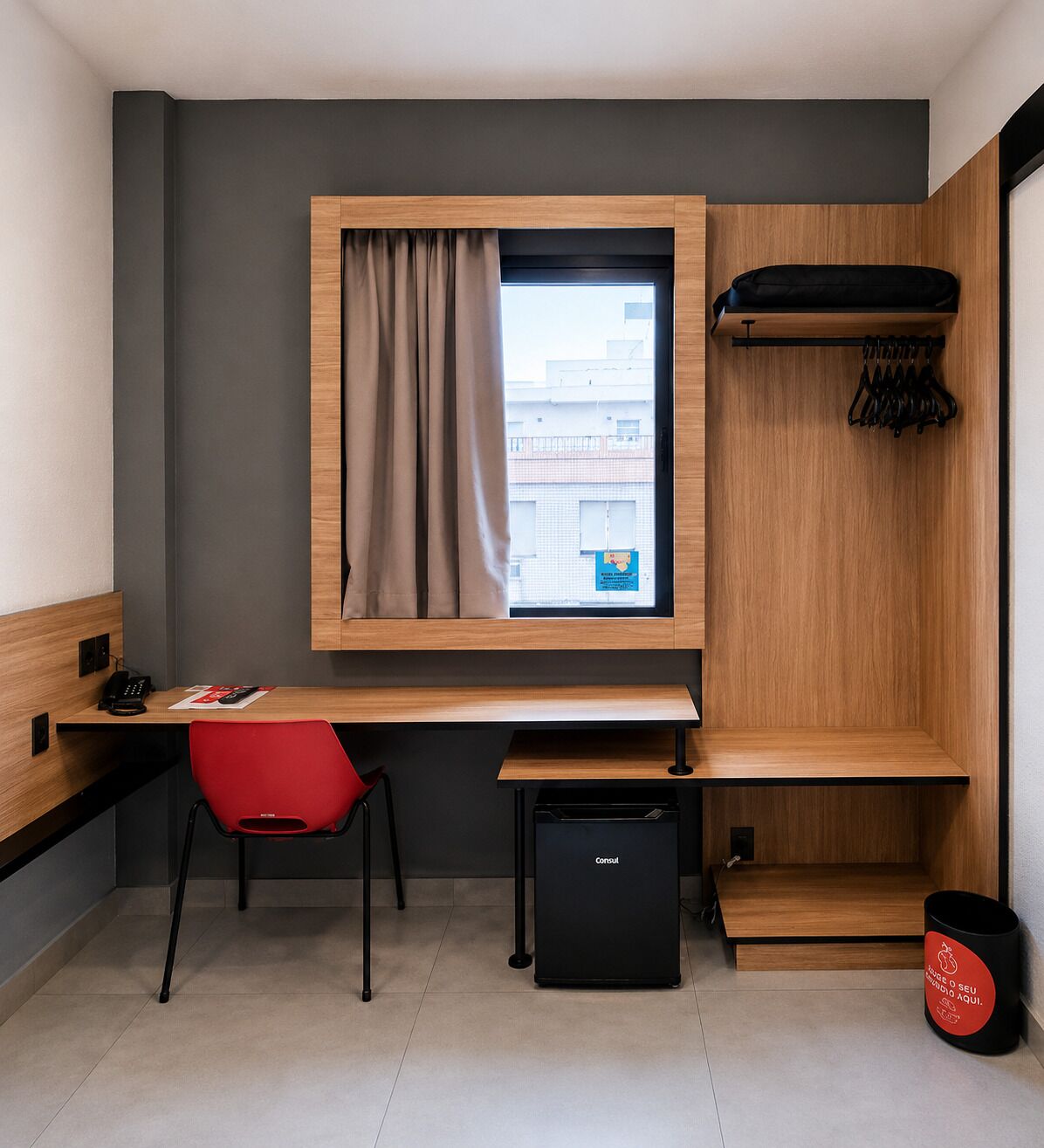 Una habitación pequeña con escritorio, una silla roja, un refrigerador negro compacto, una ventana con cortinas, un perchero y un cubo de basura.