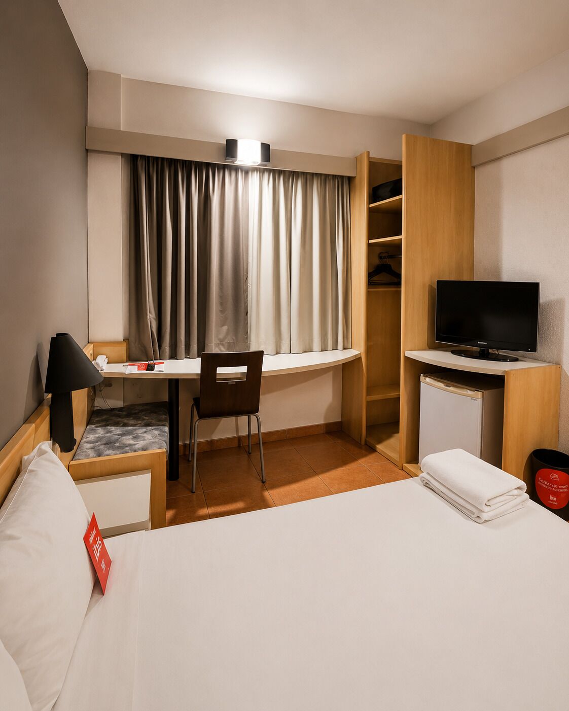 A hotel room with a bed, desk, chair, television, wardrobe, and curtains.