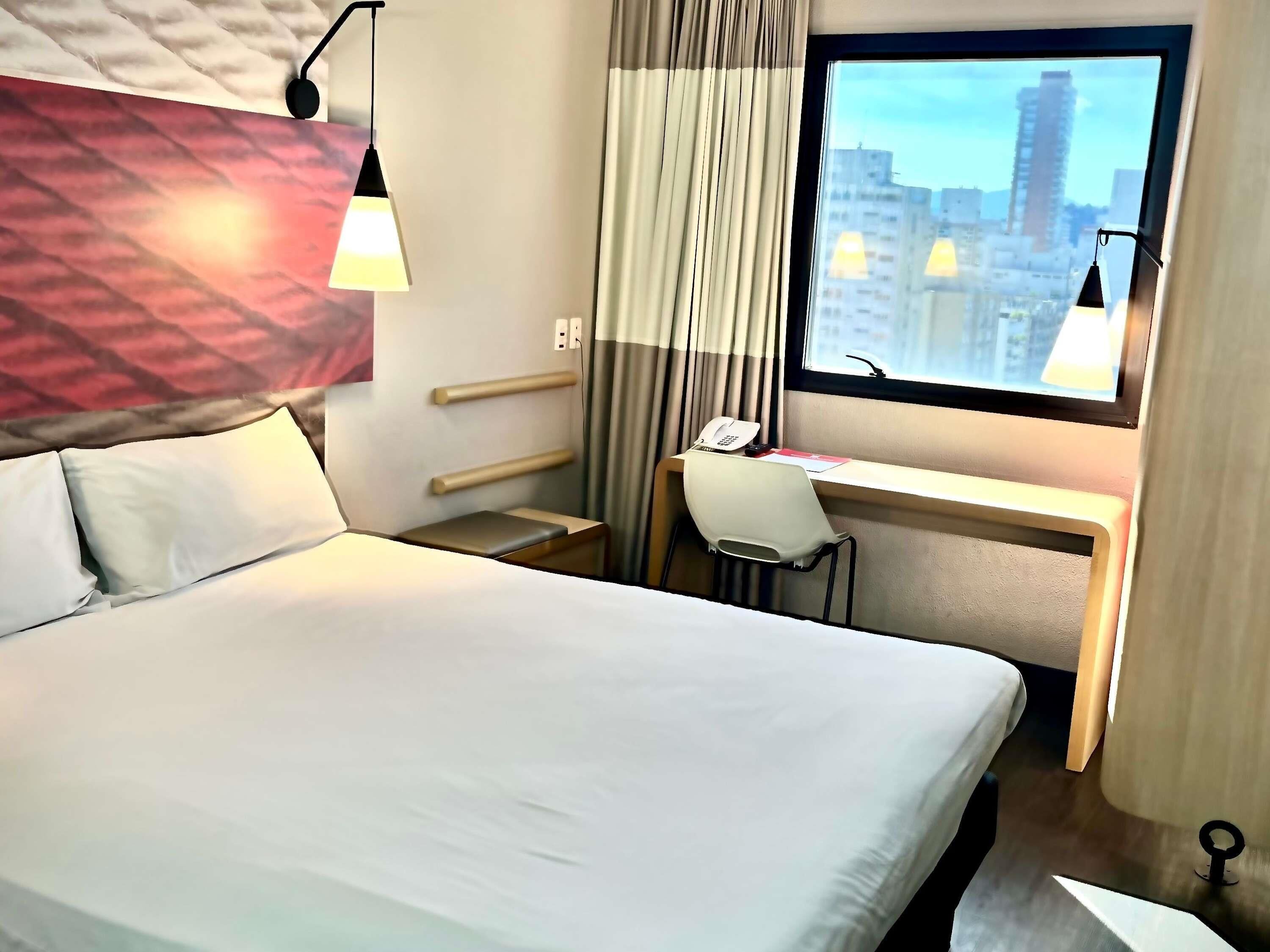 A hotel room with a bed, desk, chair, and a window with a city view.