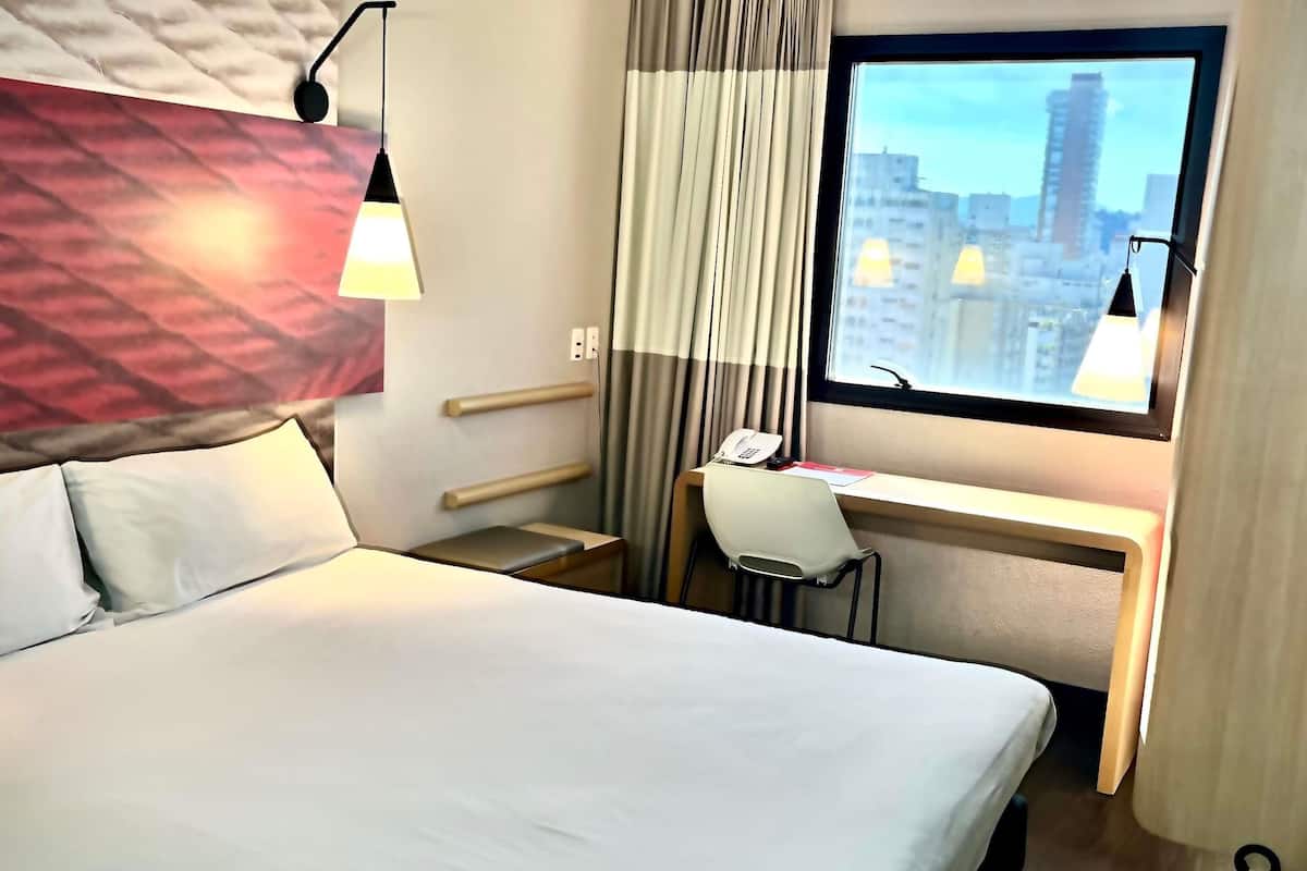 A hotel room with a bed, desk, chair, and a window with a city view.