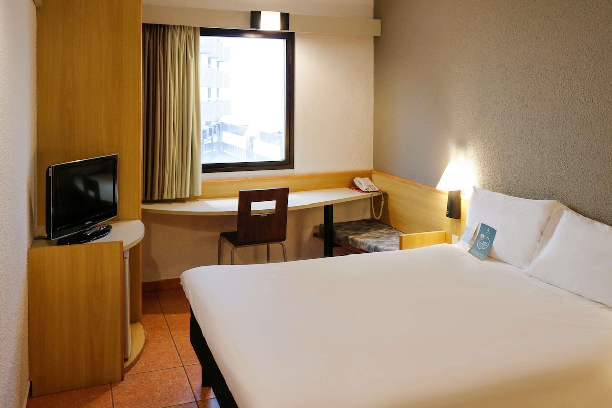 Une chambre d’hôtel comprenant un lit, un bureau avec une chaise, une télévision, une fenêtre avec des rideaux et un téléphone.