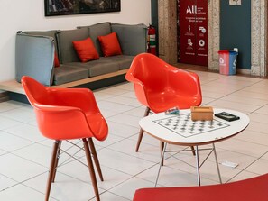 A modern living room with a grey sofa, red chairs, a small round table with a chessboard, and a fire extinguisher. - Ibis Santos Gonzaga Praia (Santos)