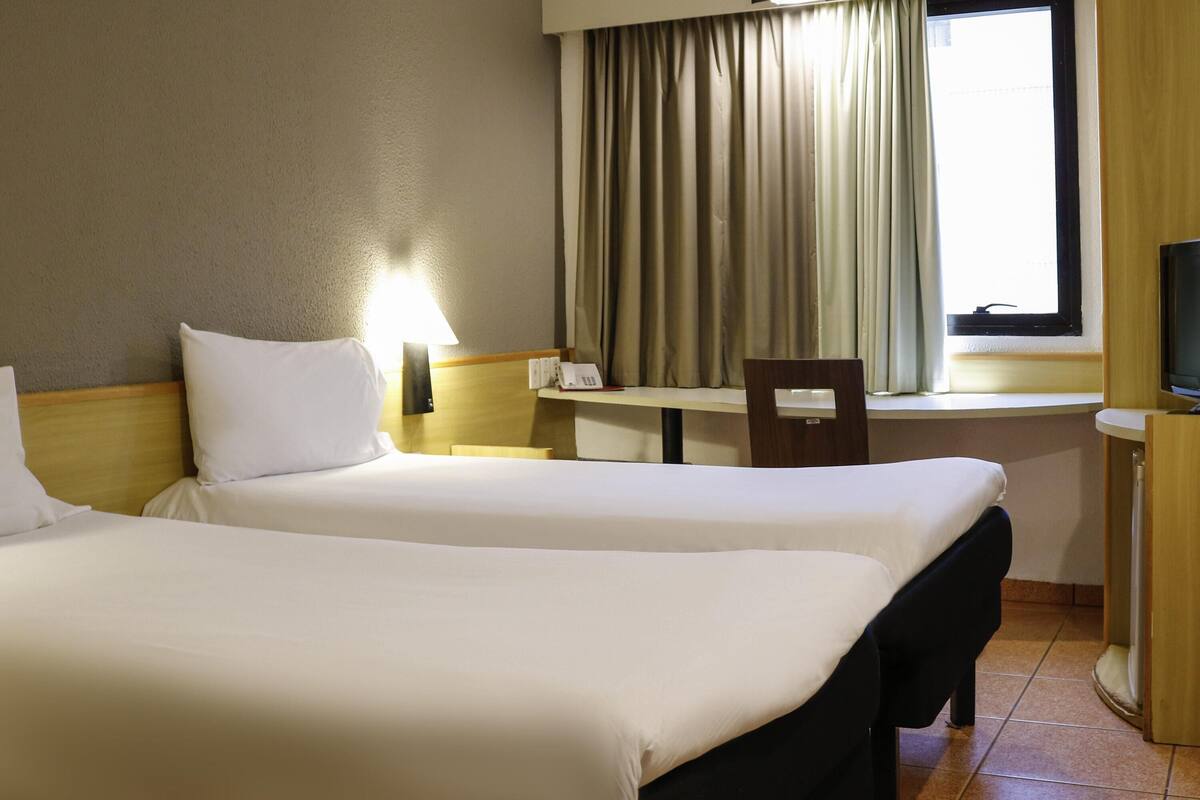 Une chambre d’hôtel avec deux lits, un bureau, une chaise, une télévision et une fenêtre avec des rideaux.