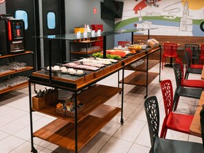 A buffet setup with various food items, a coffee machine, and red chairs. - Ibis Santos Gonzaga Praia (Santos)