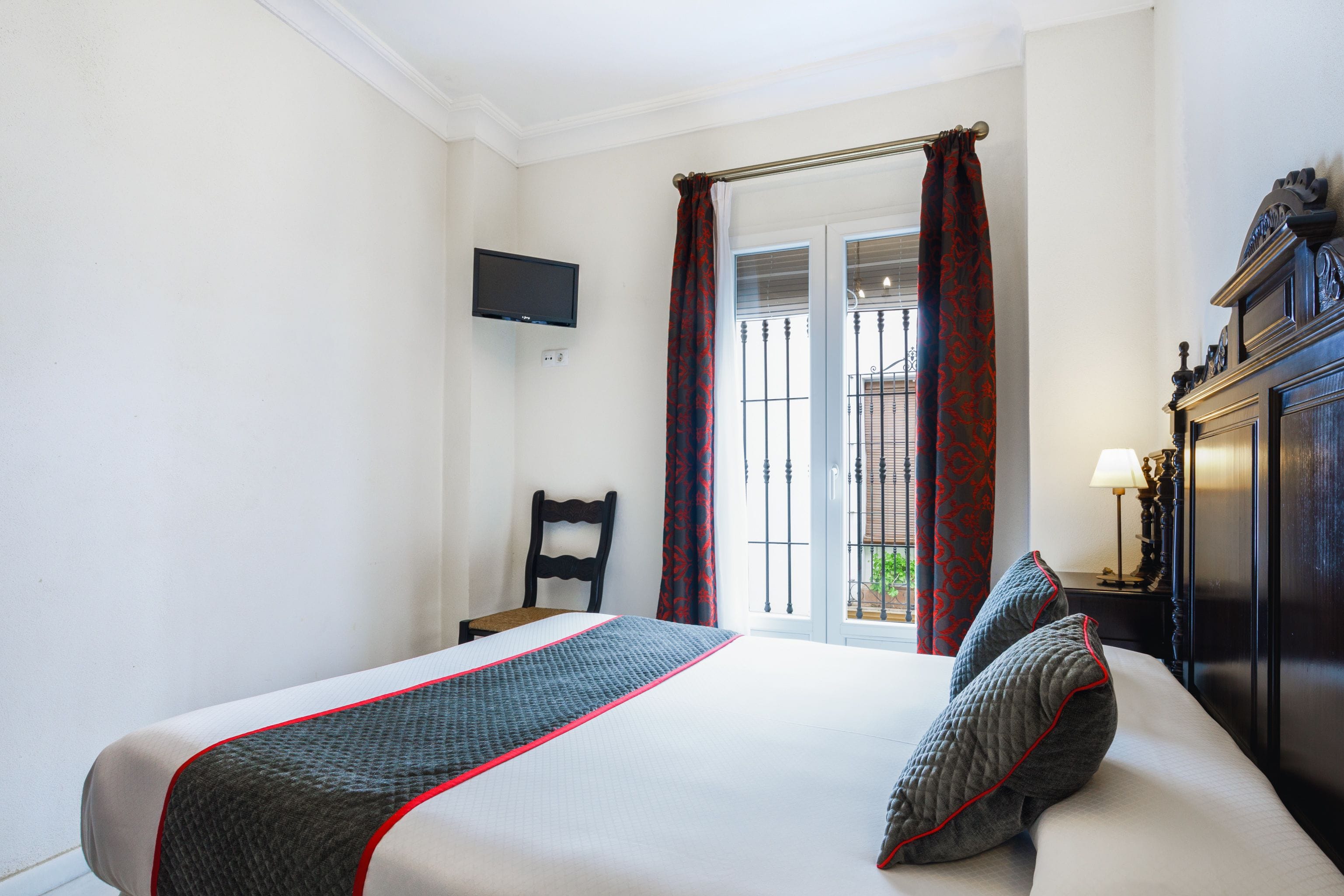 Une chambre d’hôtel avec un lit, une chaise, une télévision, une fenêtre avec des rideaux et une table de chevet avec une lampe.