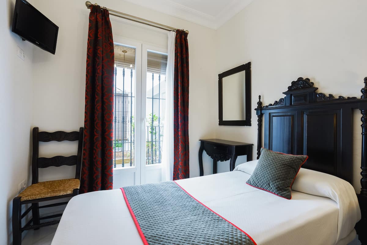 A hotel room with a bed, a chair, a small table, a mirror, and a window with curtains.