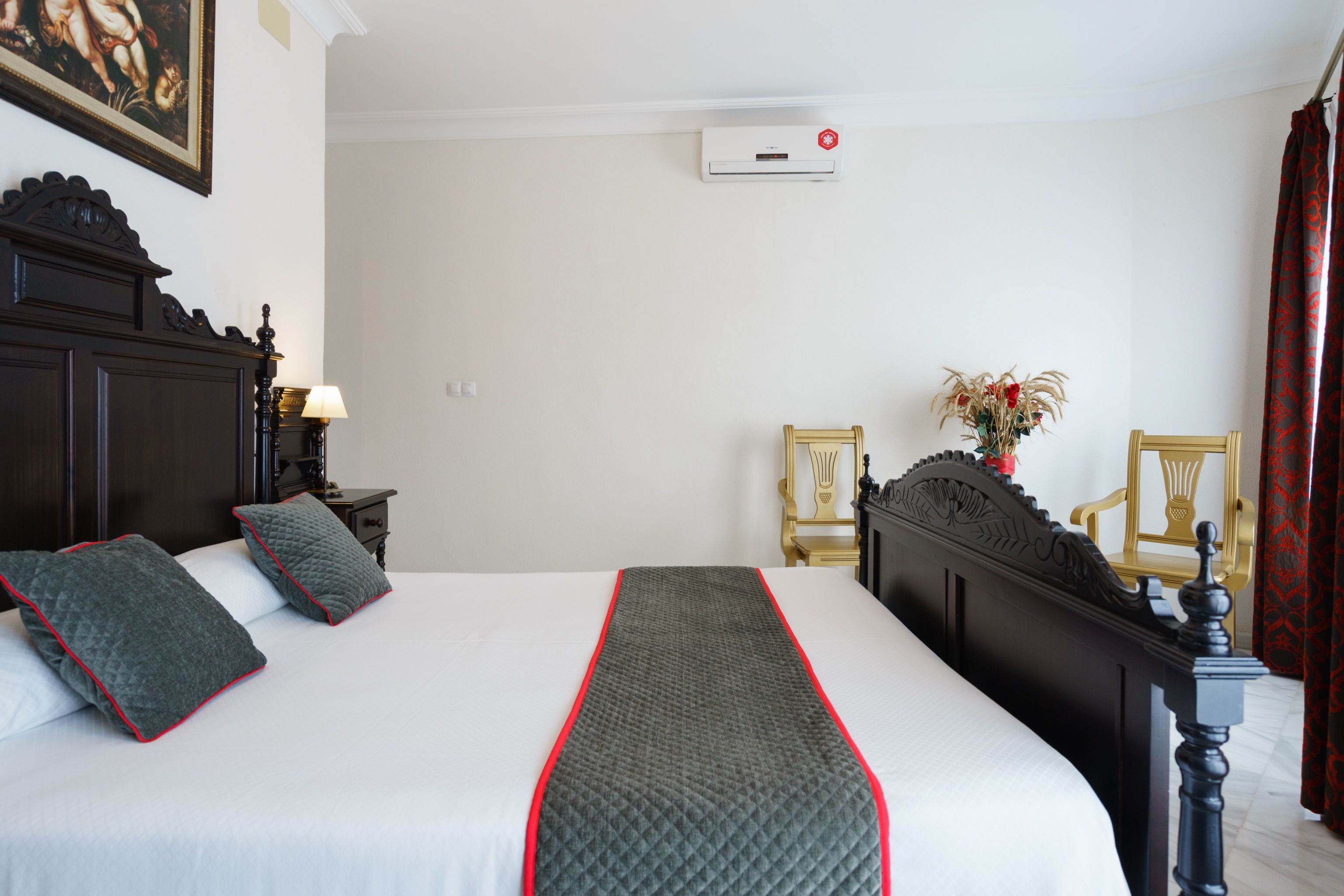 Una cama bien hecha con una colcha acolchada, dos almohadas, una mesita de noche con una lámpara, un aire acondicionado instalado en la pared y dos sillas con un jarrón de flores.