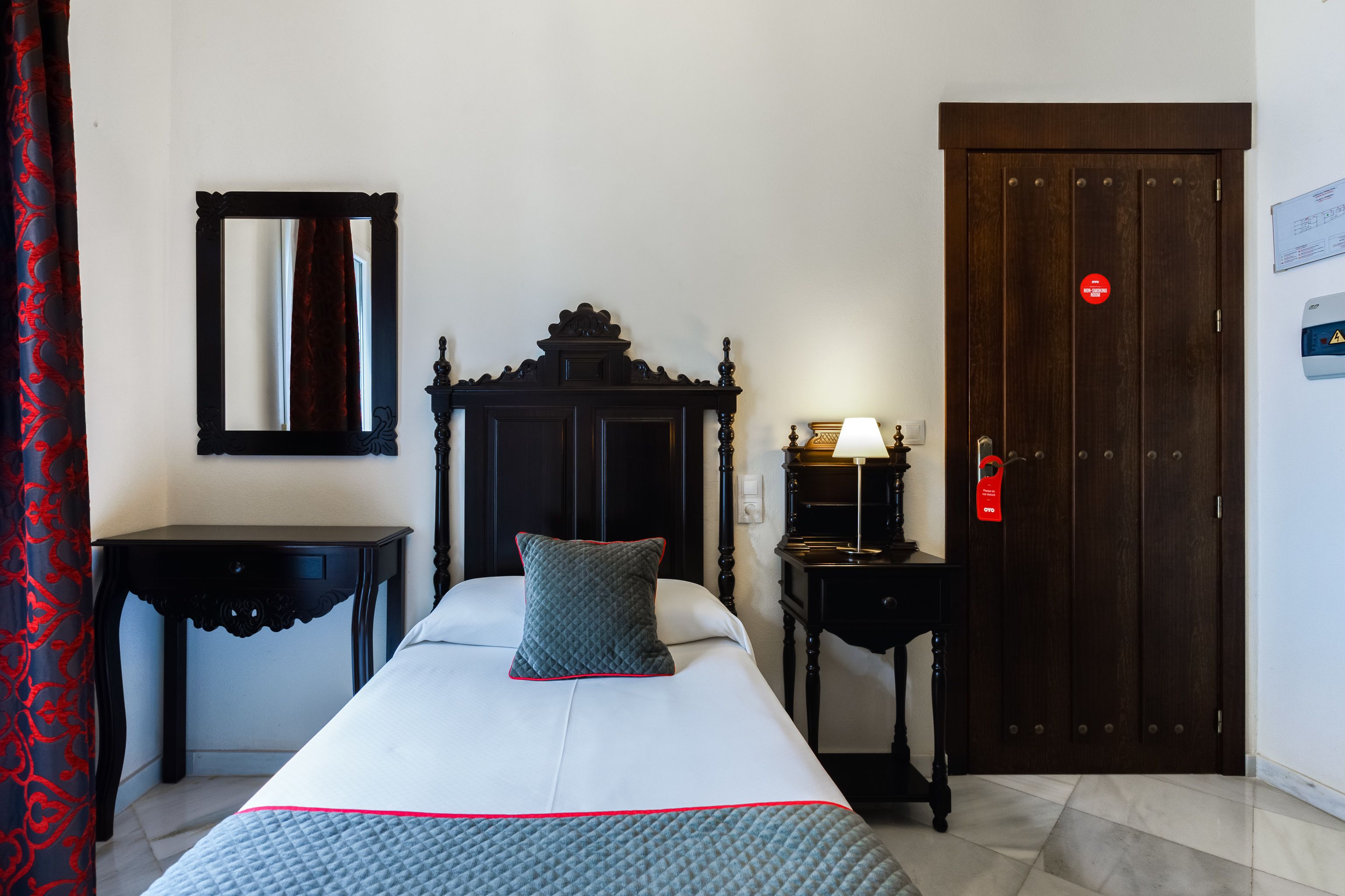 Habitación de hotel con cama, mesita de noche, cómoda, espejo, una puerta con un letrero rojo y una lámpara de pared.