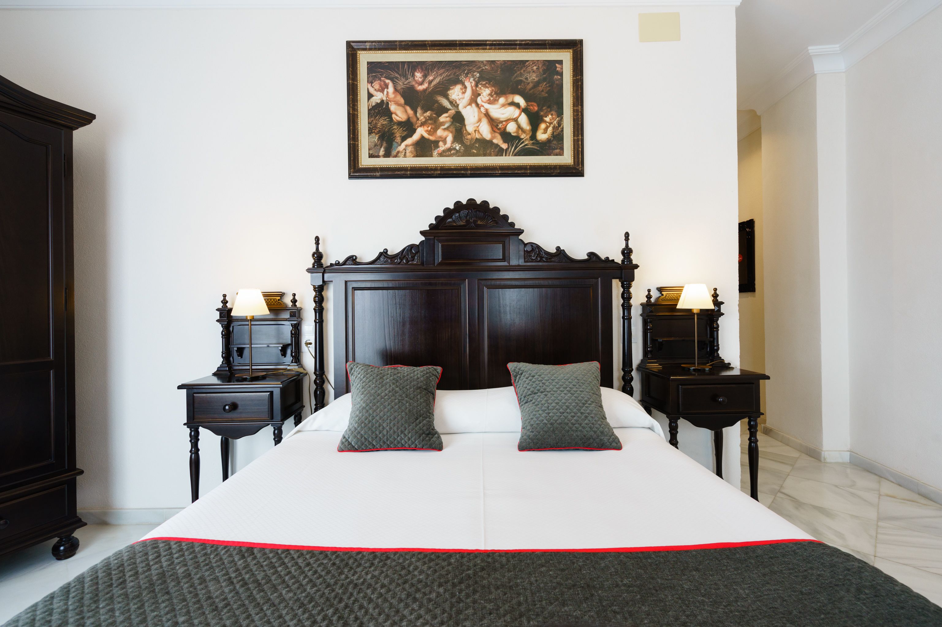 A bedroom with a large bed, dark wooden headboard, two bedside tables with lamps, a framed painting on the wall, and a dark wardrobe.