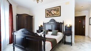Een slaapkamer met een groot bed, donker houten meubels, een kroonluchter, een schilderij aan de muur en een deur op de achtergrond.