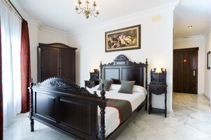 Un dormitorio con una cama grande, muebles de madera oscura, una araña, un cuadro en la pared y una puerta al fondo.