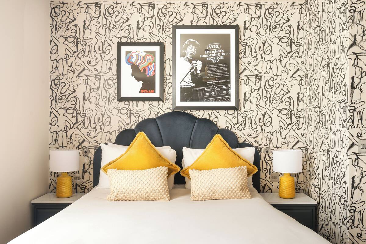 A hotel room with a patterned wallpaper, a bed with yellow and white pillows, and two framed pictures on the wall.