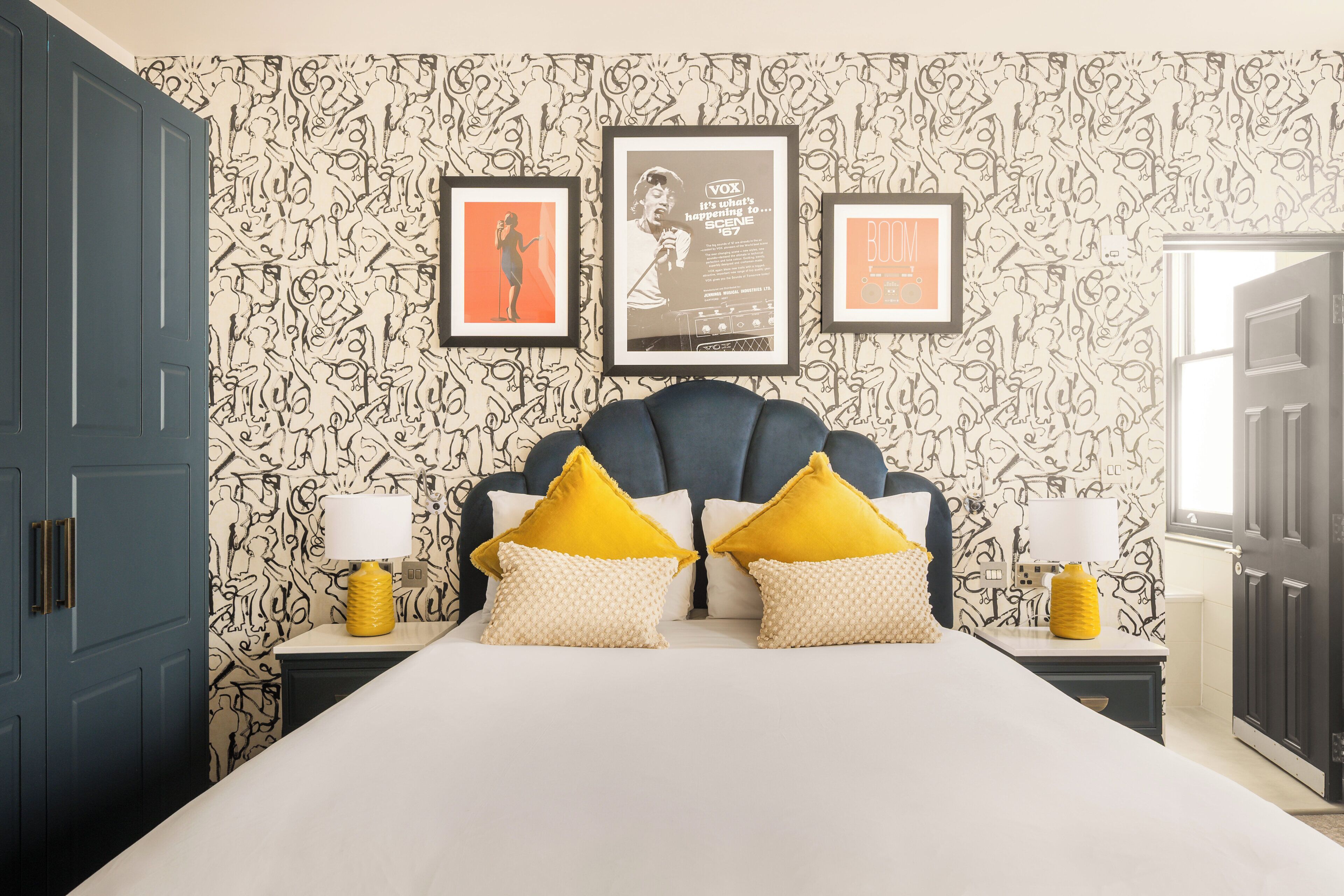 A bedroom with a bed, two nightstands, a headboard, and a wall with patterned wallpaper and framed posters.