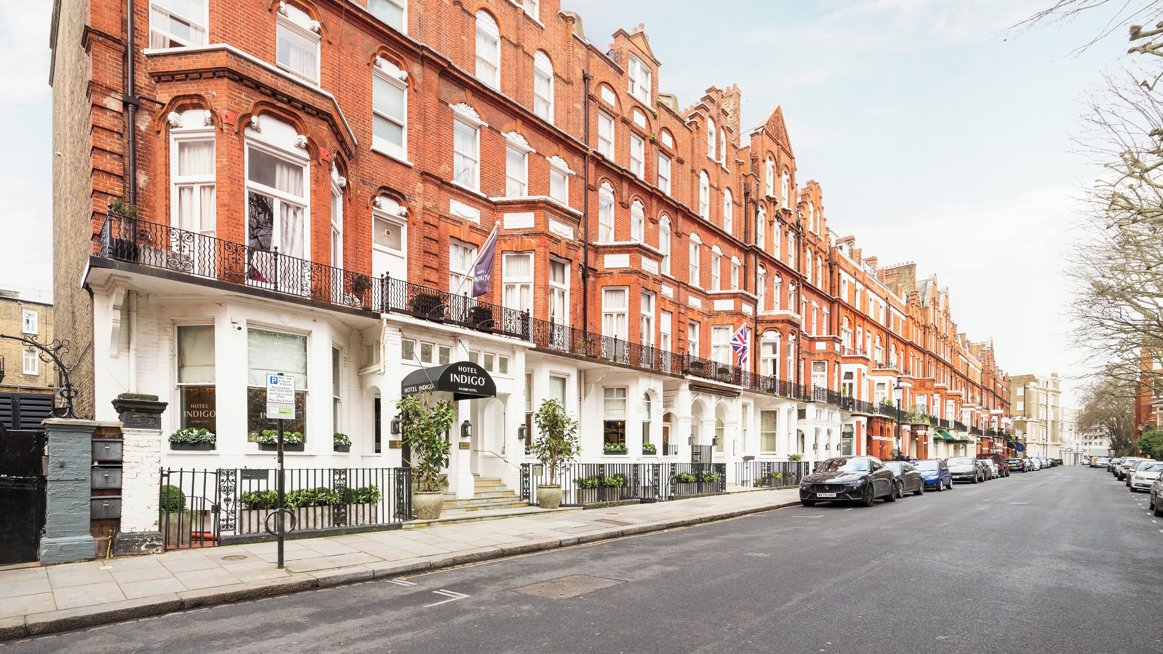 A row of red-brick terraced houses with white facades, a black metal fence, and a street with parked cars.