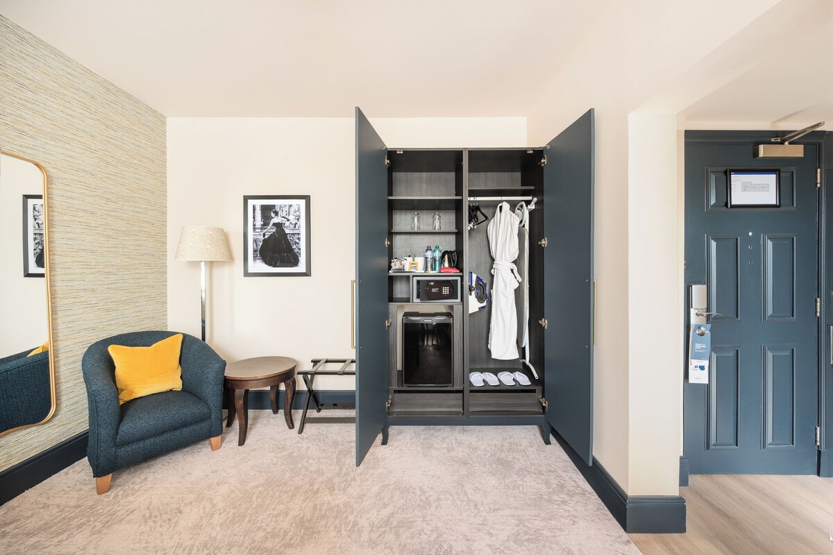 A hotel room with a dark blue door, a small seating area with a yellow cushion, a framed picture on the wall, and a closet with a robe and slippers.