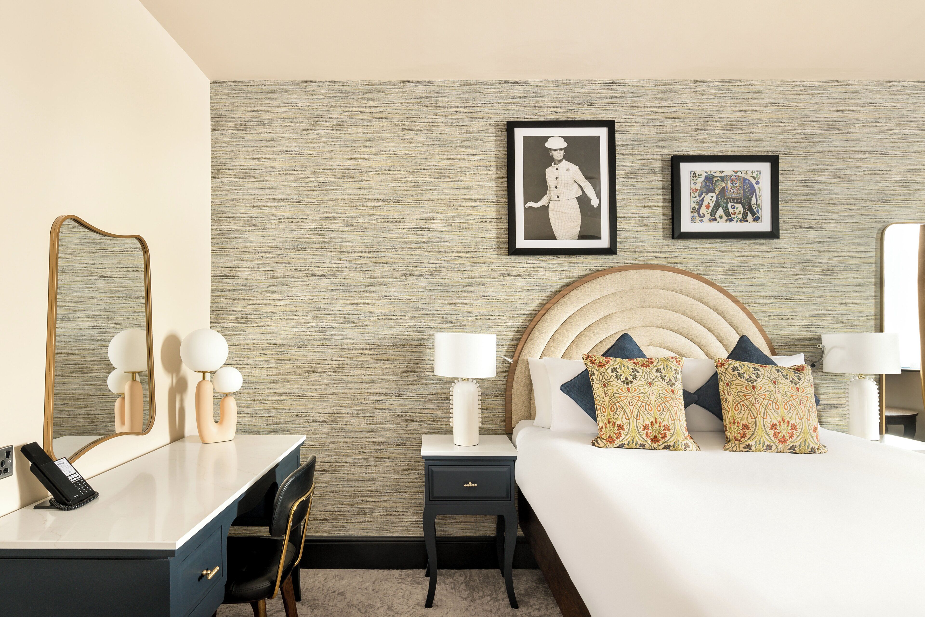A hotel room with a bed, bedside tables, a desk, a chair, a mirror, and two framed pictures on the wall.