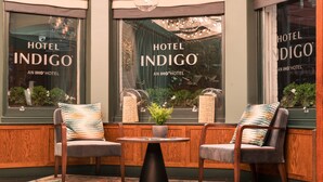 Un área de estar con dos sillas, una mesa, una planta en maceta y un ventanal con el logotipo de Hotel Indigo.