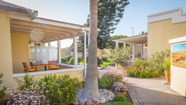 Un porche cubierto con una mesa y sillas, rodeado de un jardín ajardinado con diversas plantas y una palmera.