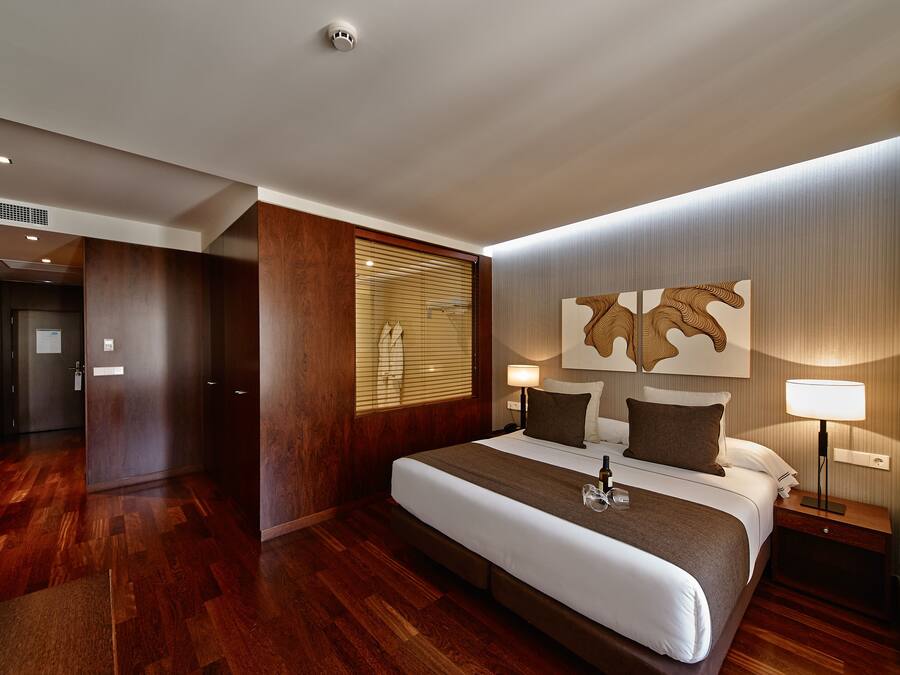 A modern hotel room with a large bed, wooden flooring, a wooden partition, a wall-mounted artwork, and a bedside table with a lamp.