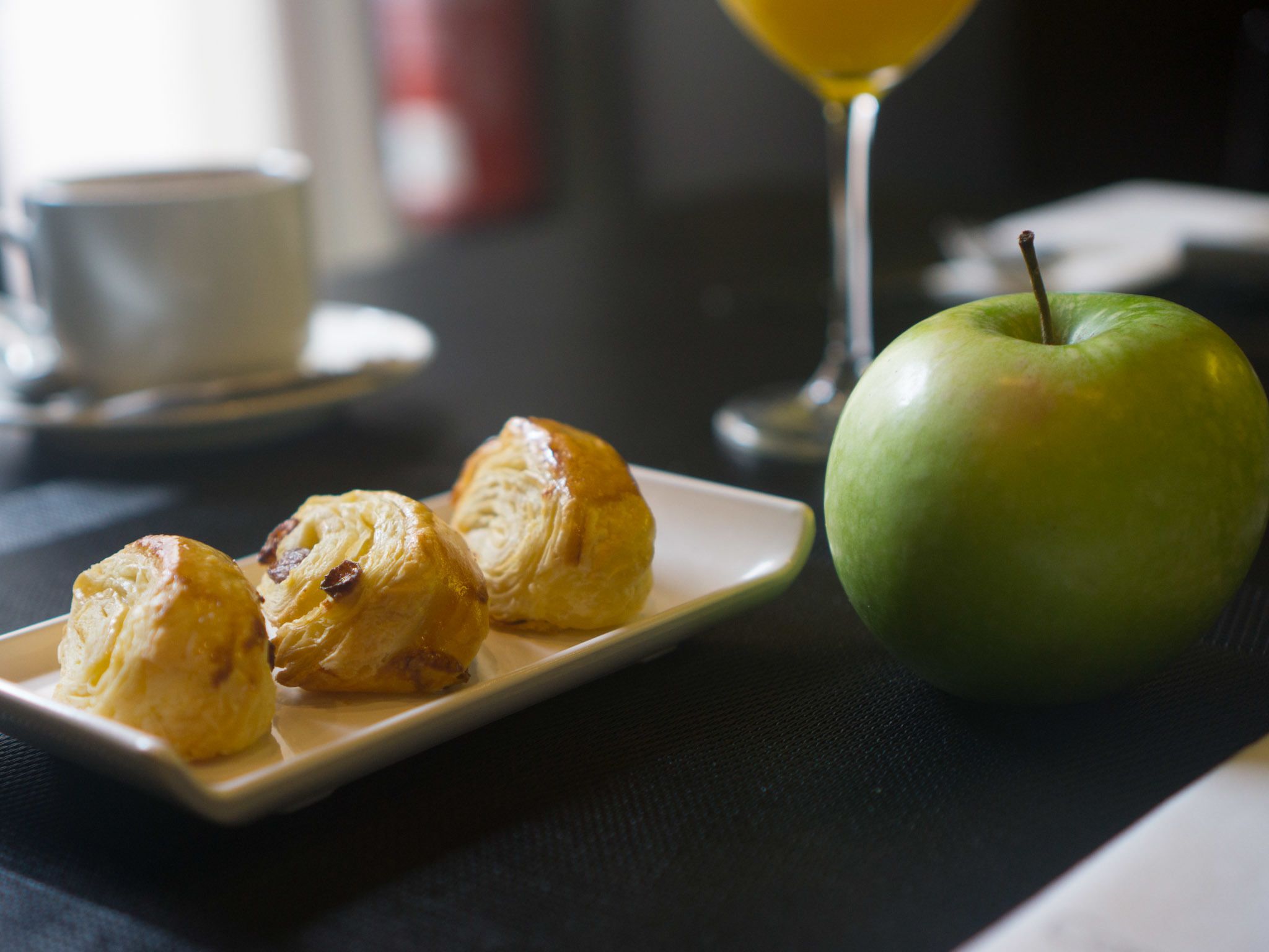 Un plato con pasteles y una manzana verde sobre una mesa, con un vaso de jugo de naranja y una taza al fondo.