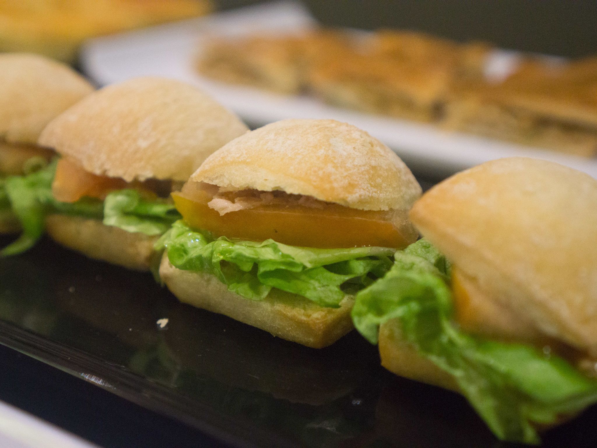 A row of sandwiches with lettuce and a slice of tomato on a black tray.