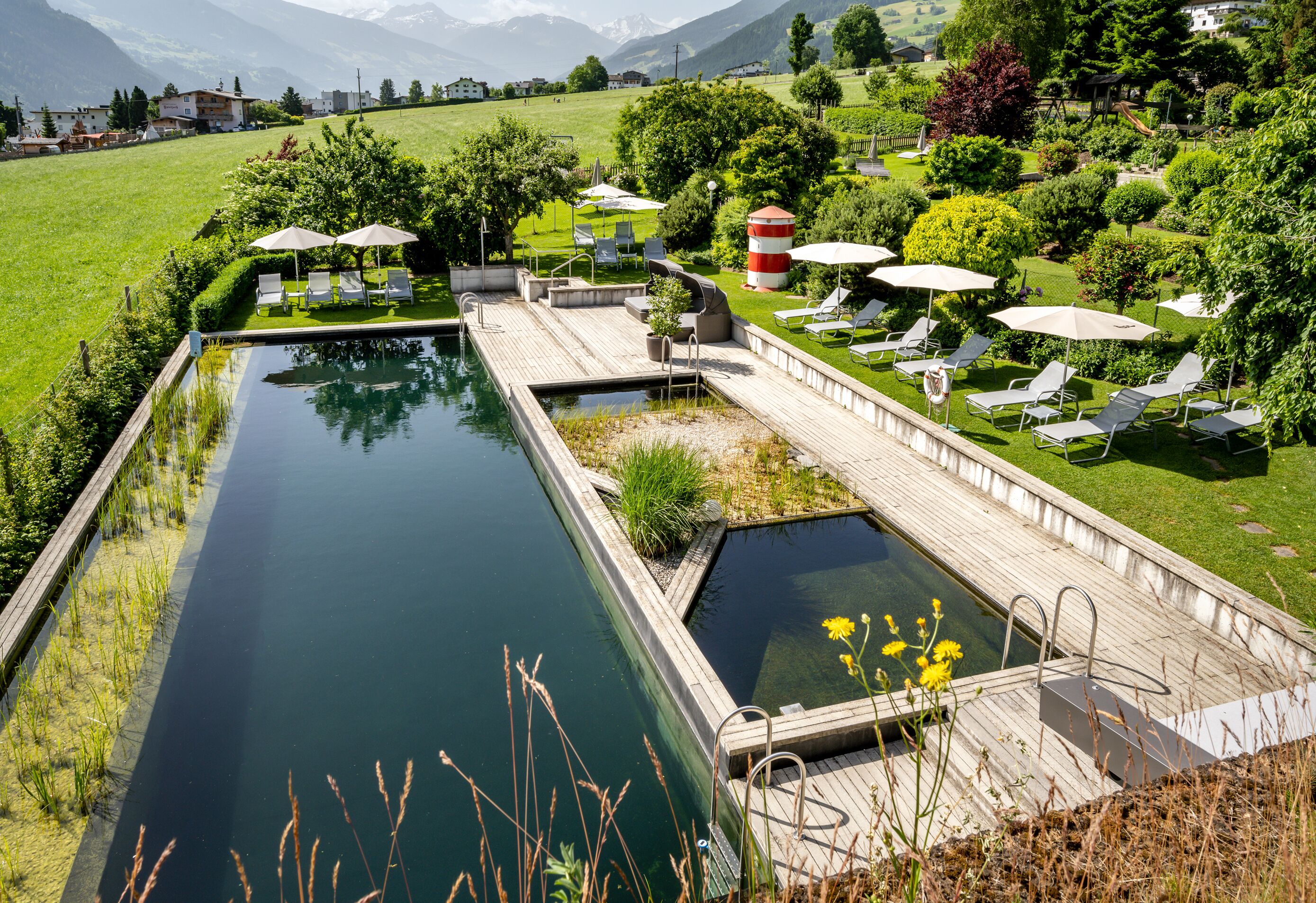 Ein ruhiger Poolbereich mit Holzterrasse, Liegestühlen und Sonnenschirmen, umgeben von üppiger Vegetation und einer Bergkulisse.