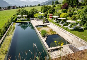 A serene pool area with a wooden deck, lounge chairs, and umbrellas, surrounded by lush greenery and a mountain backdrop.
