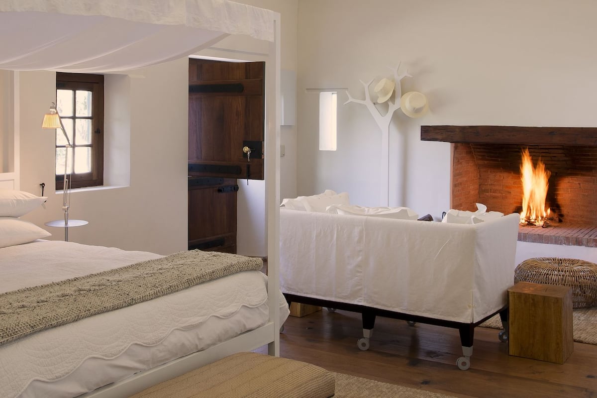 A bedroom with a four-poster bed, a bedside table, a chair, a fireplace, and a framed botanical print on the wall.
